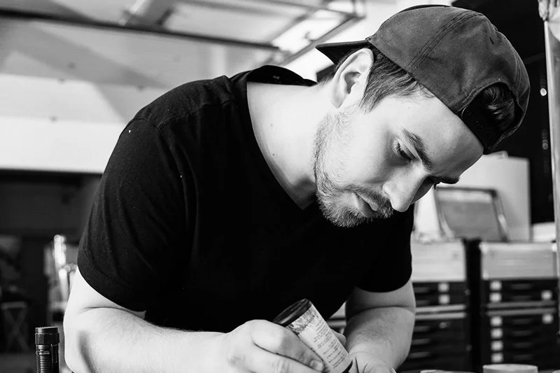 Melvin Klietzing A young man wearing a backward baseball cap and a black T-shirt leaning over a table, opening a can in a workshop or garage setting.