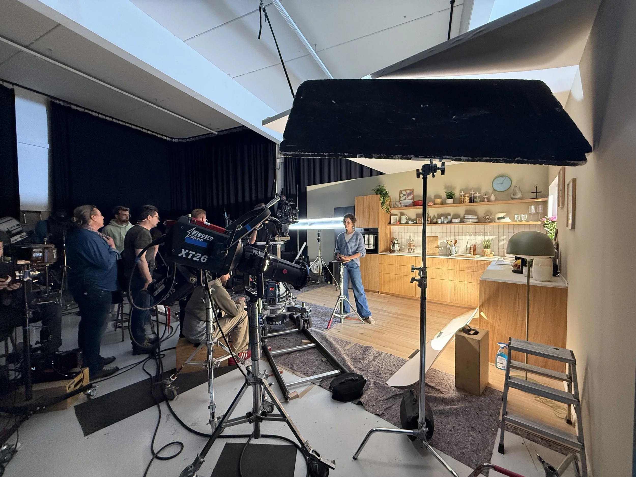 A filming crew setting up for a shoot in a kitchen studio. The scene includes a camera operator, multiple crew members, lighting equipment, and a woman standing in front of a kitchen set with wooden cabinets and open shelves, decorated with various k