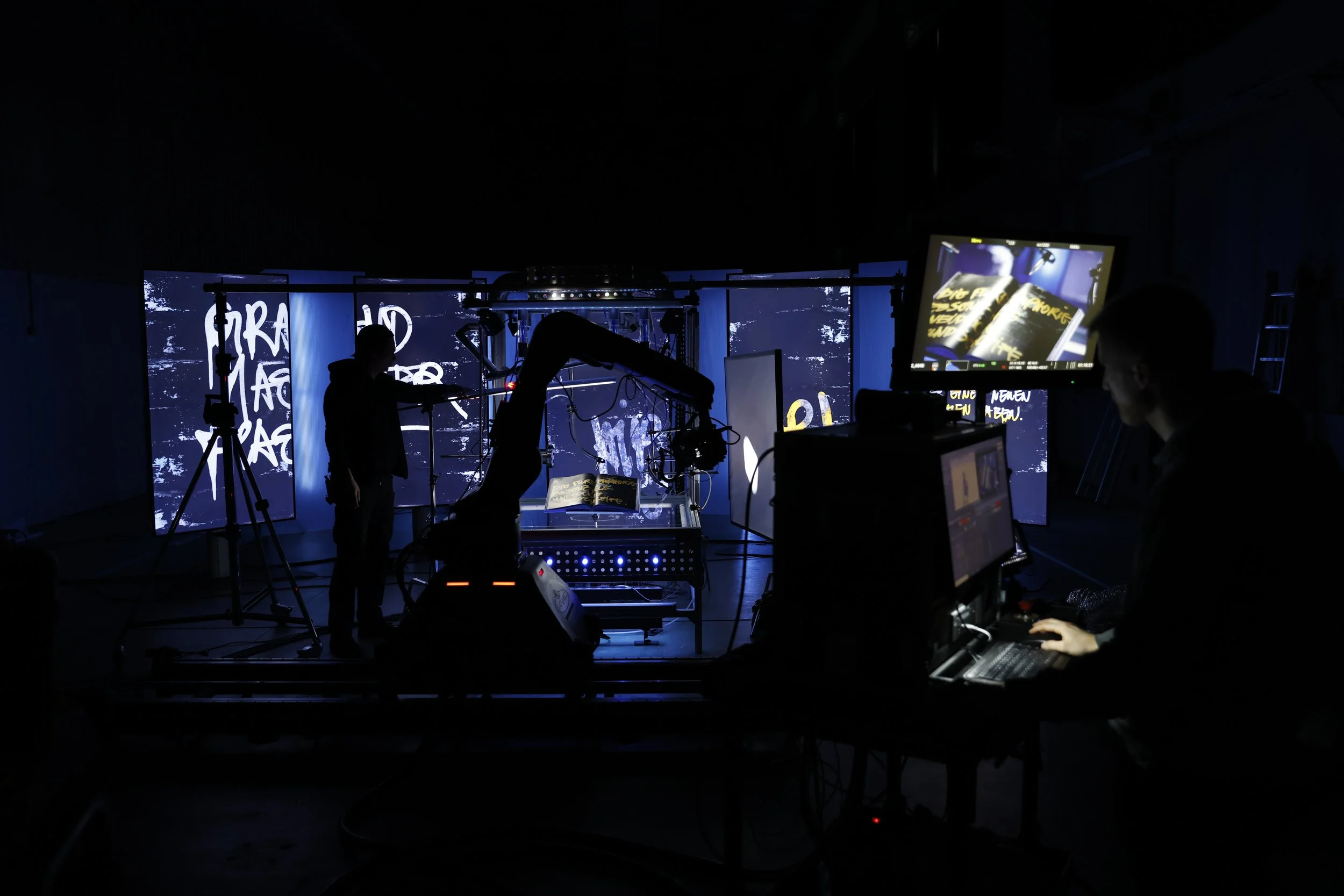 A dark studio with a robotic arm, camera, and multiple screens showing graffiti-style text and visuals, as crew members work on a production or filming setup.