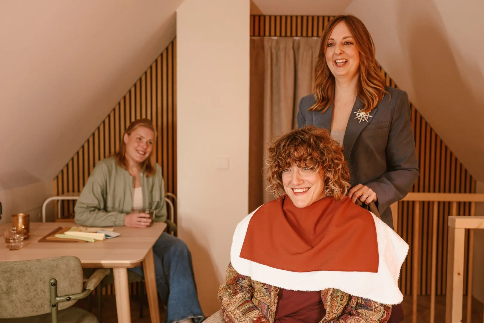 Vrouw krijgt een kleurdoek om haar schouders terwijl ze lachend voor de spiegel zit. Op de achtergrond zit een vrouw met een drankje aan een tafel.