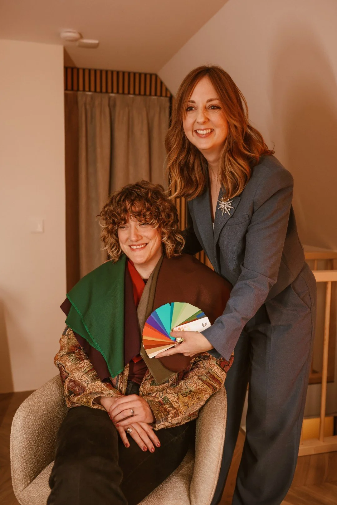 Twee vrouwen lachen en poseren voor de camera in een interieur. Een vrouw zit in een stoel en houdt een kleurenwaaier vast, terwijl de andere vrouw naast haar staat en haar handen op haar schouders heeft. Ze dragen stijlvolle kleding en de sfeer is vrolijk.
