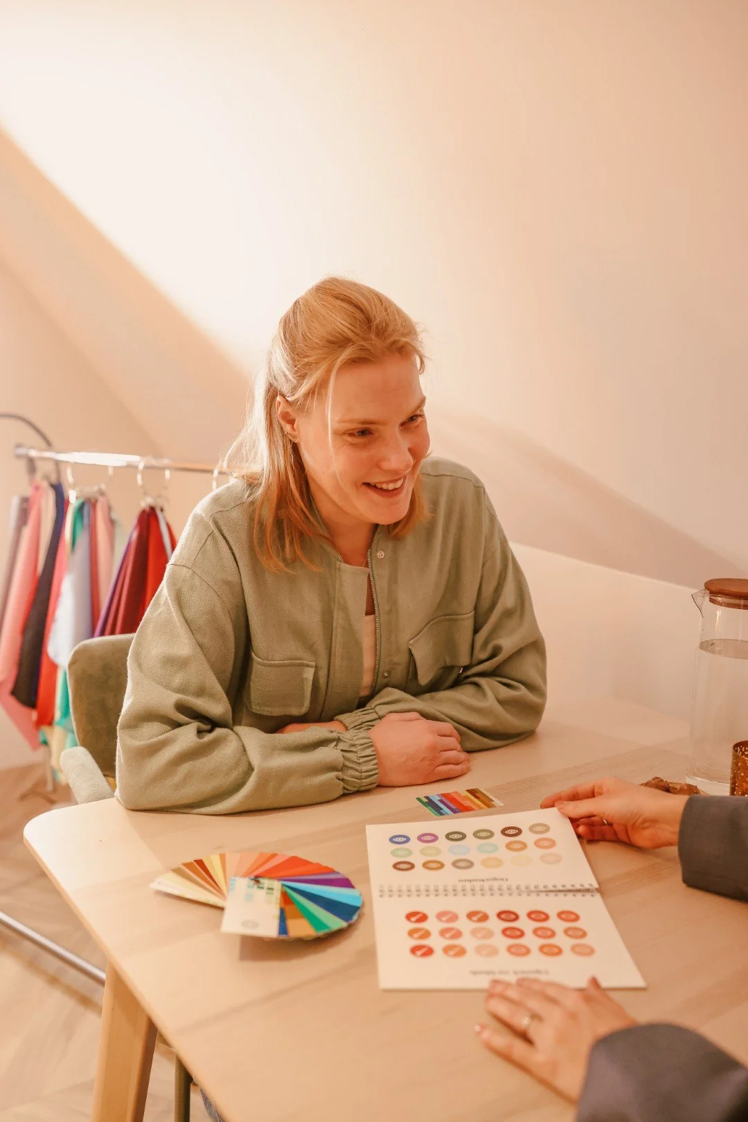 Vrouw zit aan tafel tijdens een gesprek, met kleurkaarten en een kleurenadviesboek voor zich, in een lichte kamer met kledingrekken op de achtergrond.