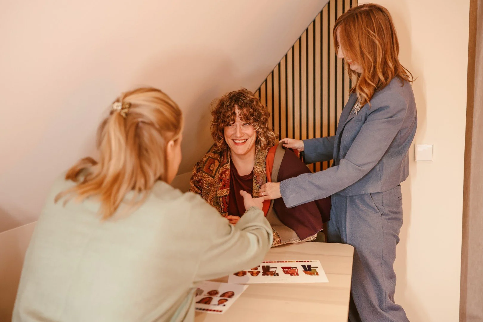 Twee vrouwen helpen een vrouw met een lach op haar gezicht met haar jas. Ze zitten aan een tafel met foto''s op papier.