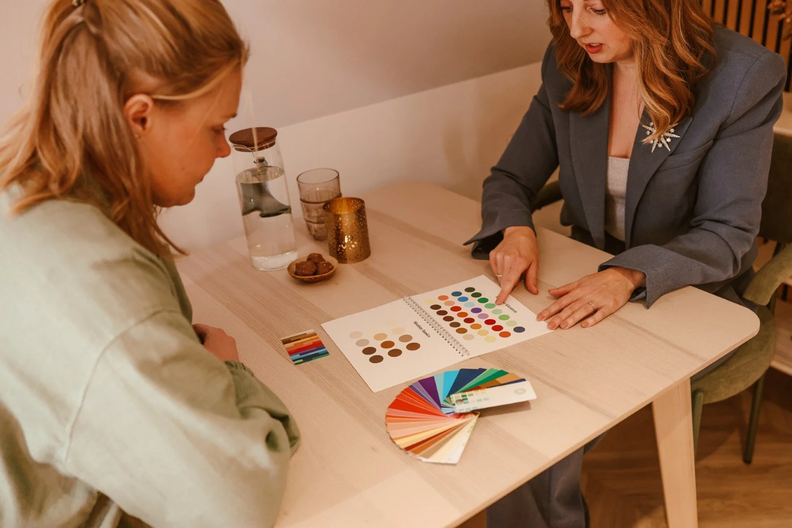 Twee vrouwen die een kleurpalet en een kleurenschema bekijken aan een tafel. Op de tafel liggen kleurenstaaltjes, een kleurwaaier en een kleurkaart.