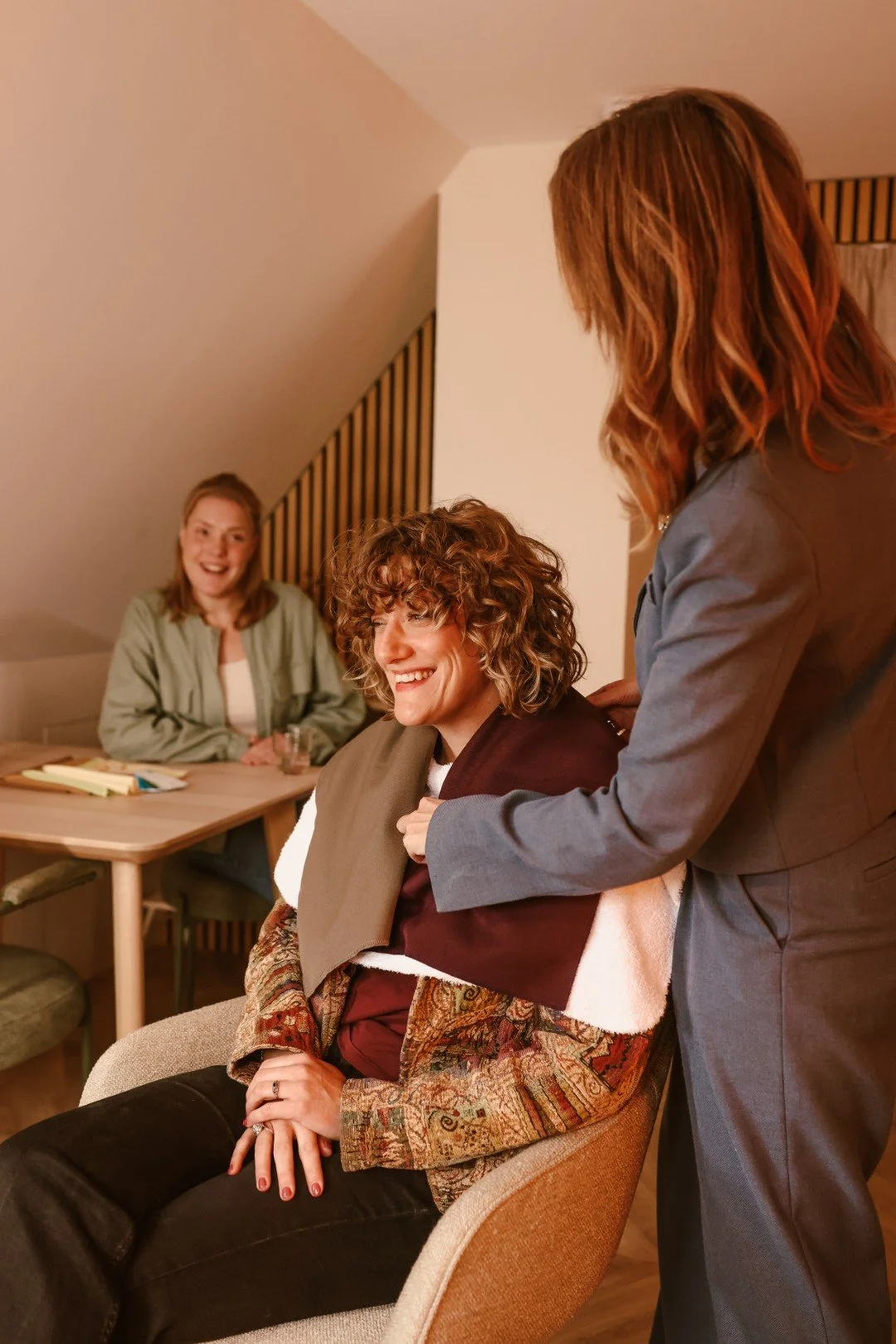 Twee vrouwen zitten samen met een lachende vrouw die wordt geknipt door een haarstylist in een gezellige kamer.