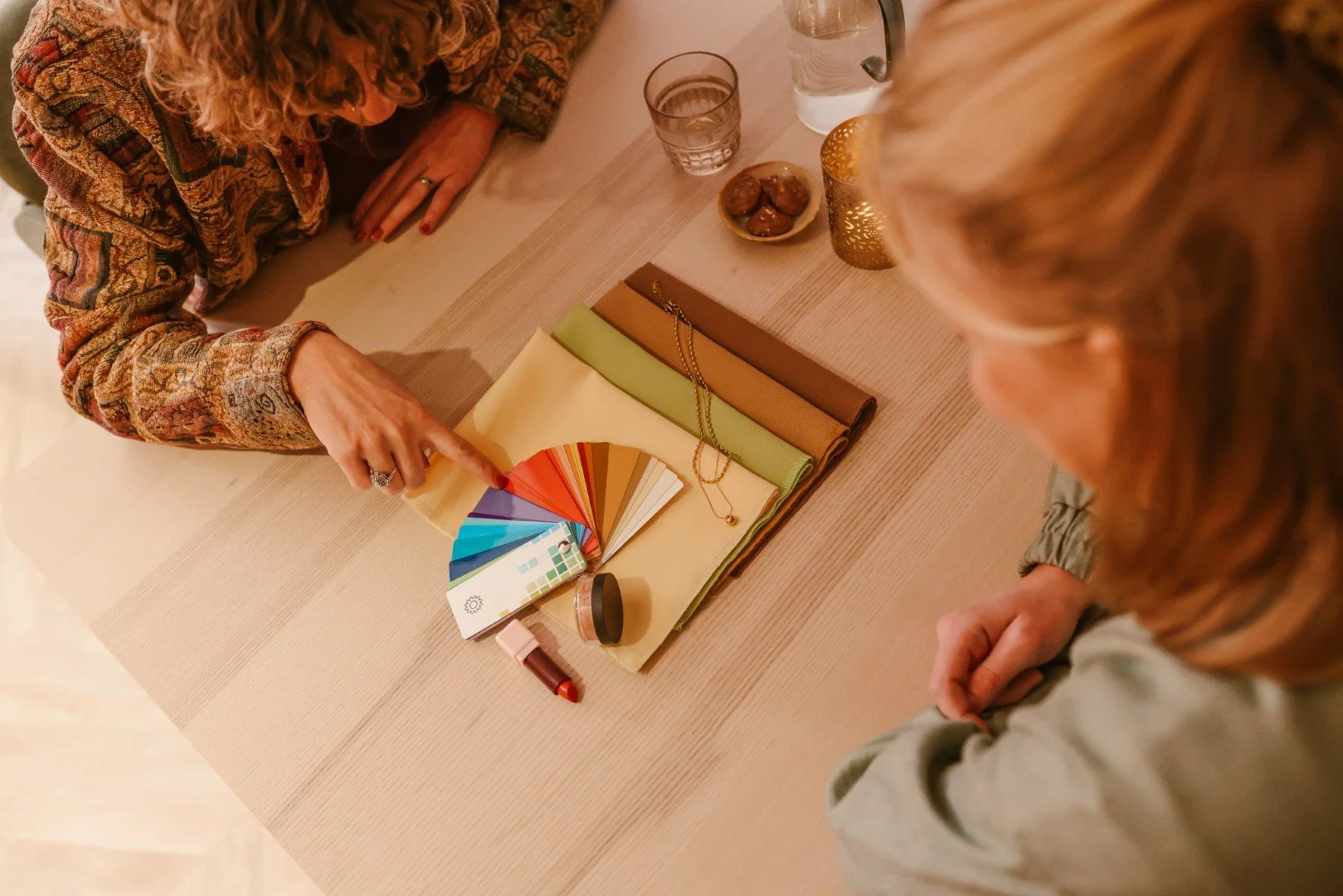 Twee vrouwen bespreken kleuren en stoffen bij een tafel met kleurkaarten, stoffenstalen en accessoires.