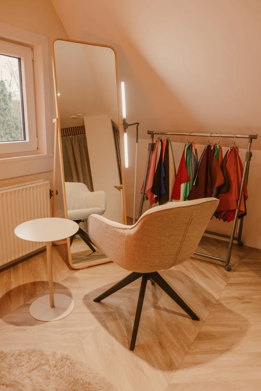Een spiegel met een houten rand, een fauteuil, een kleine witte tafel, een kledingrek met kleurrijke stoffen en een raam in een kamer met houten vloer.