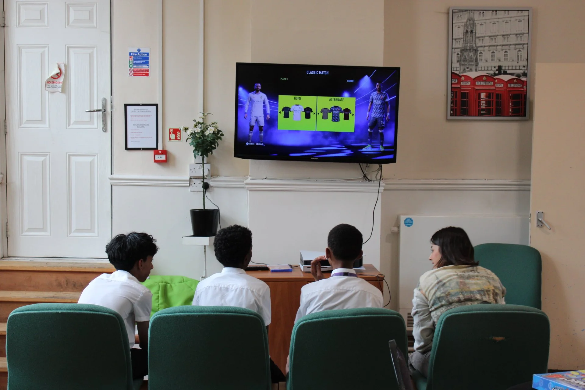 A group of students sitting in chairs watching a soccer video on a television screen. The screen displays a soccer game selection menu with options for team jerseys and uniform designs.
