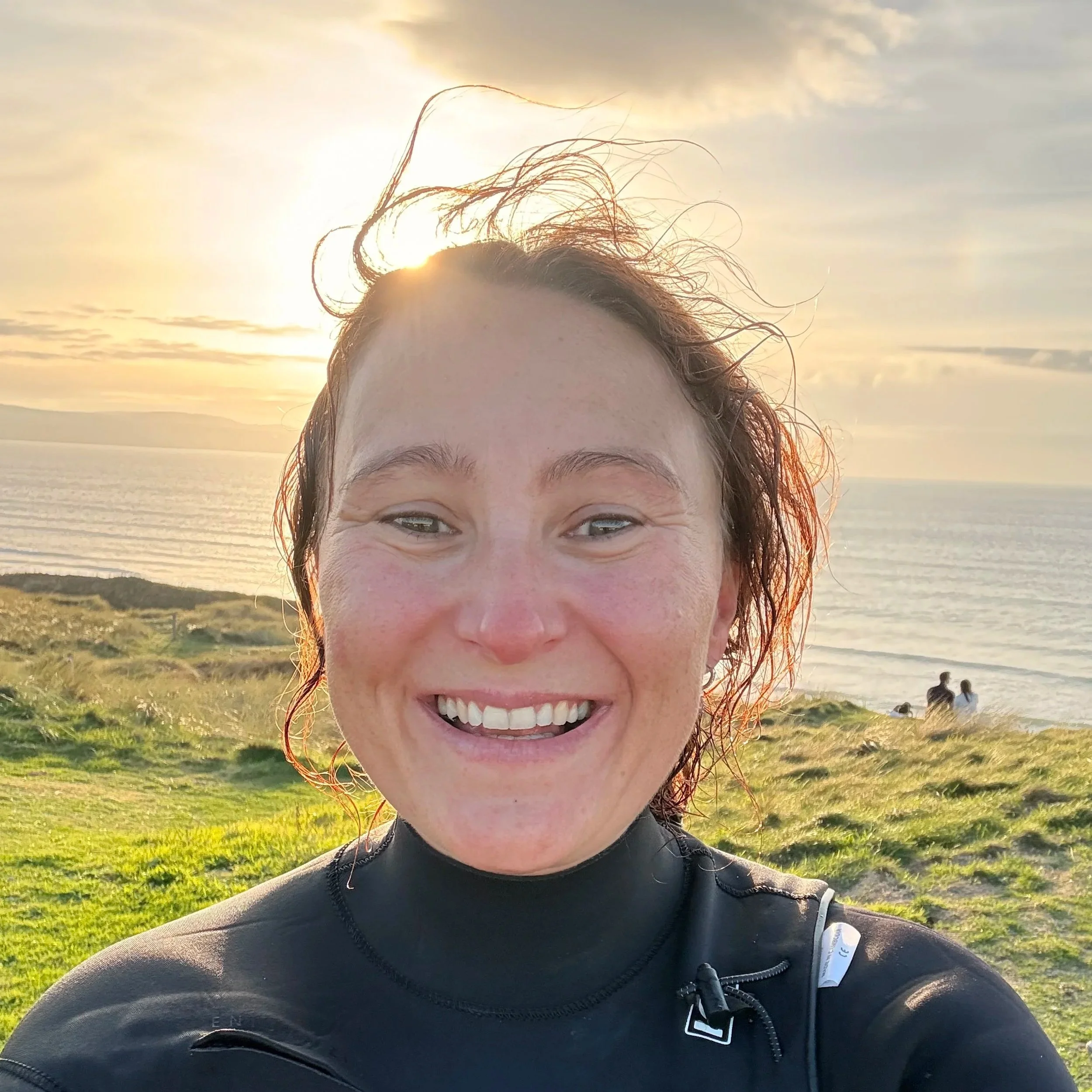 A woman smiling outdoors at sunset with ocean and grassy landscape in the background.