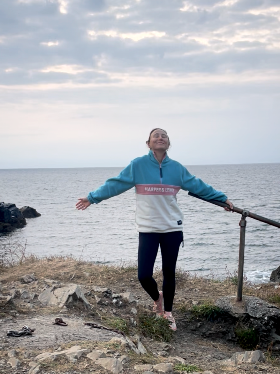 A woman standing on a rocky coastal area with her arms outstretched and eyes closed, smiling, near the ocean during cloudy weather.