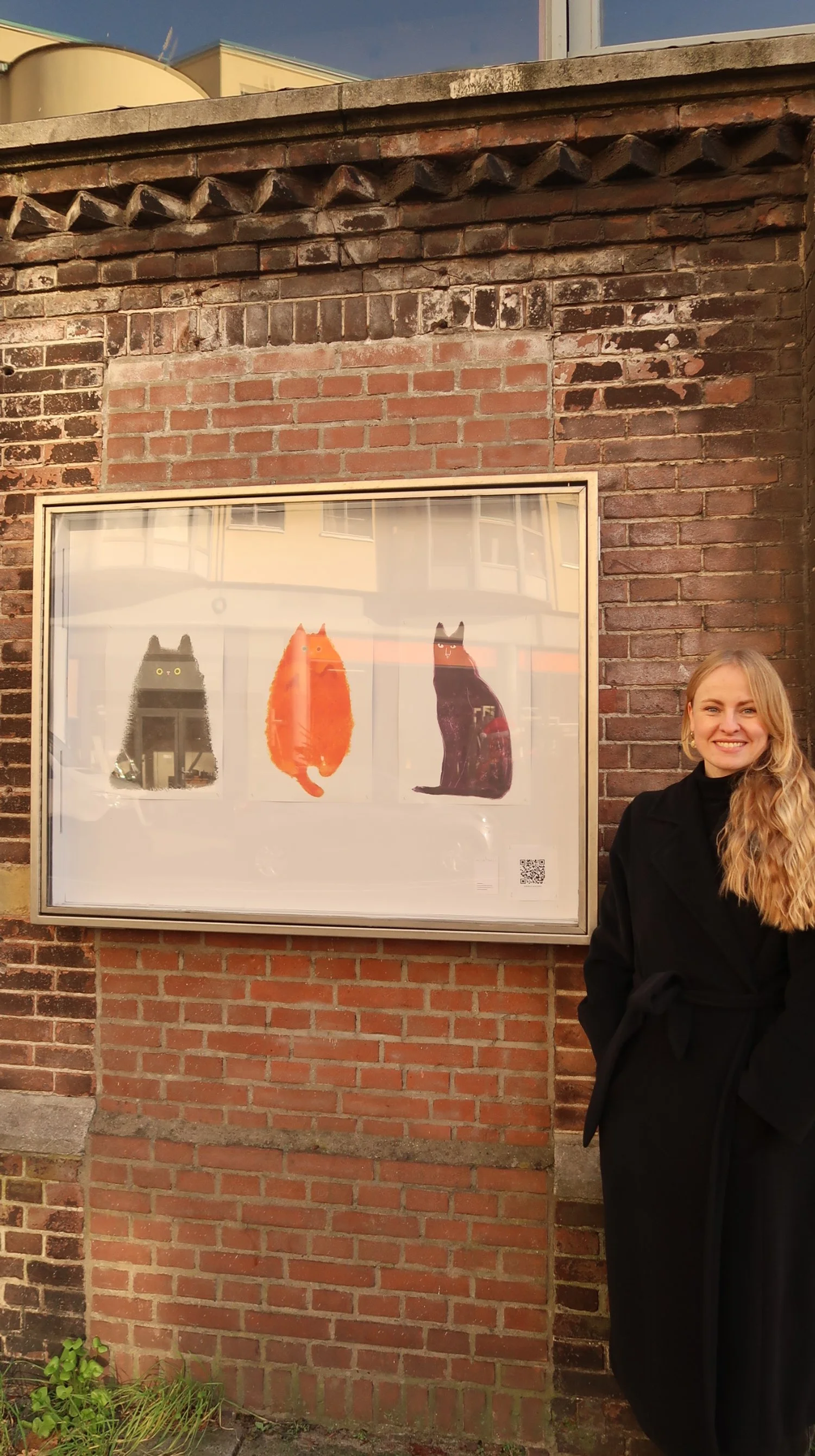 A woman standing next to a framed poster of three illustrated cats on a brick wall.