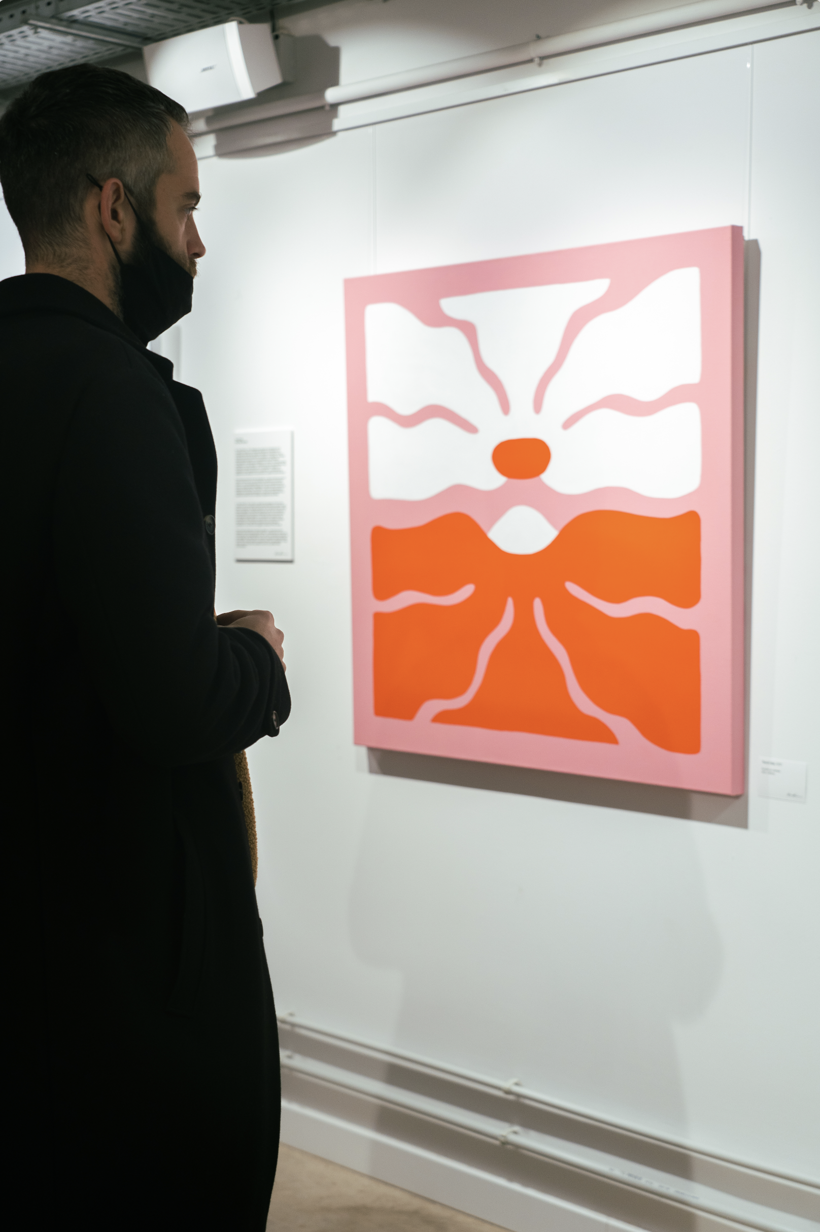 A person wearing a black coat and a black face mask looking at an abstract pink and orange painting in an art gallery.