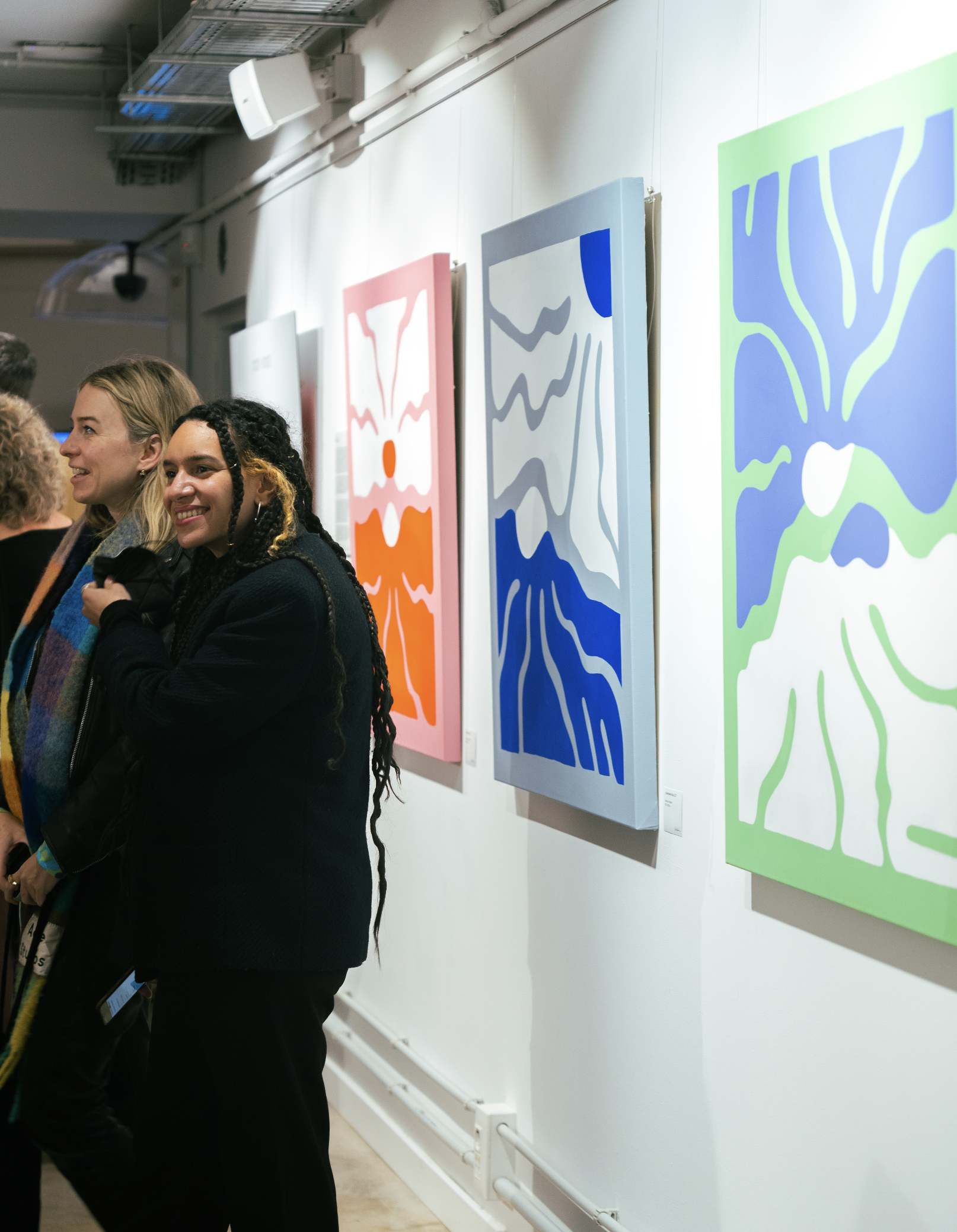 People attending an art gallery, looking at colorful abstract paintings hung on a white wall.