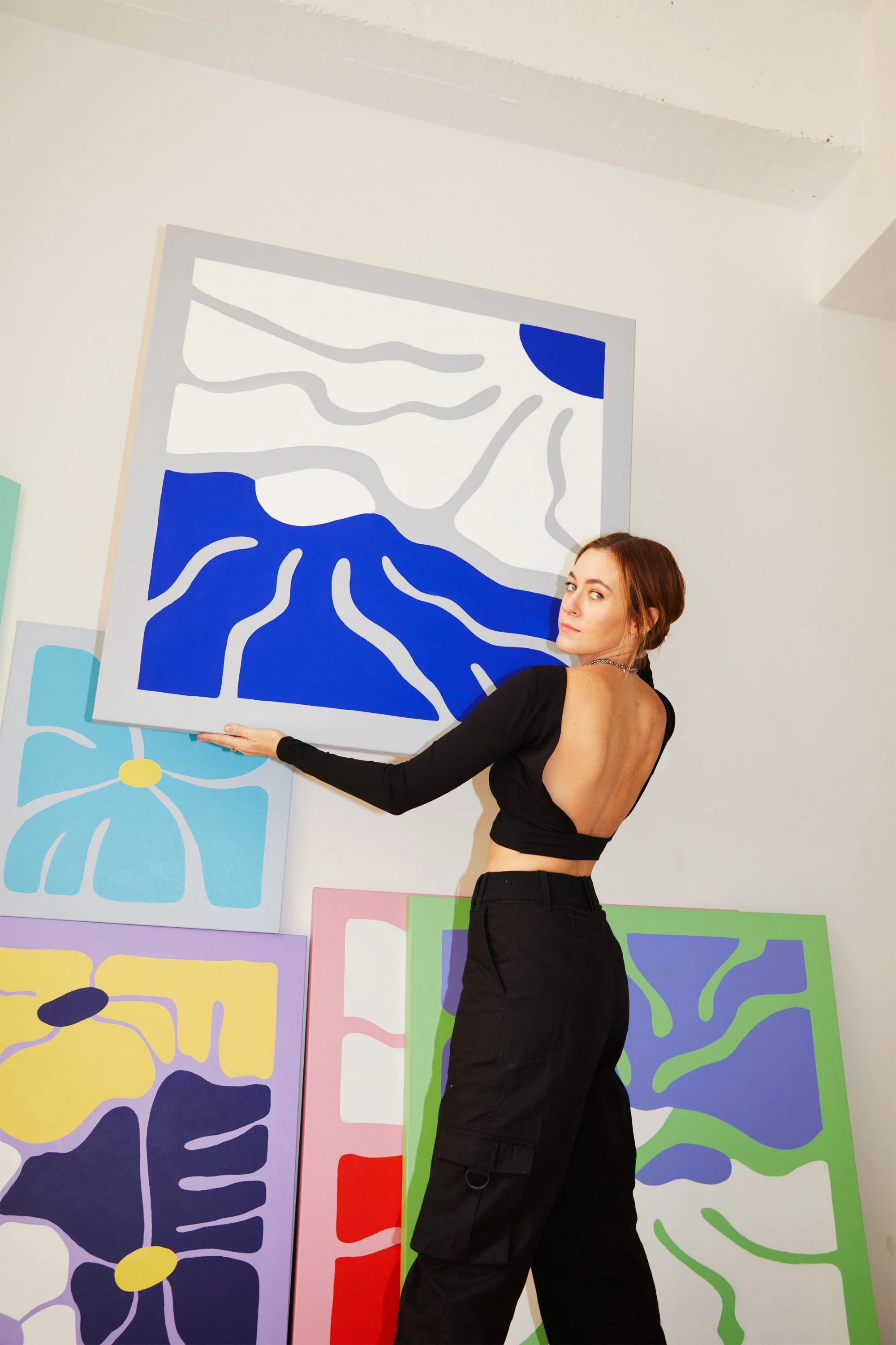 A woman with brown hair standing next to a modern abstract painting with blue, white, and gray colors. Several other colorful abstract paintings are visible in the background.
