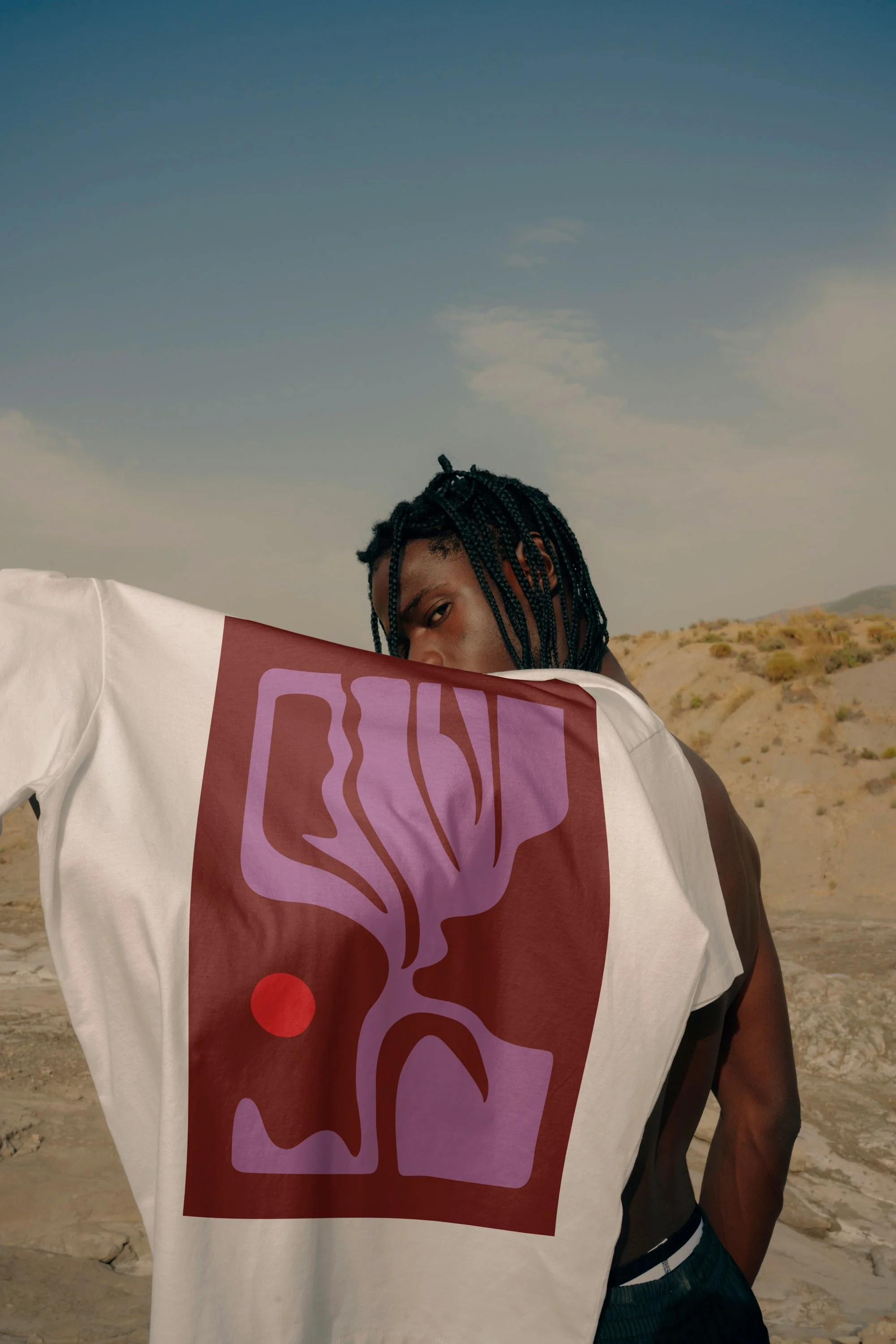 A man with braided hair standing in a desert landscape, holding a white t-shirt with a purple and red abstract design partially covering his face.