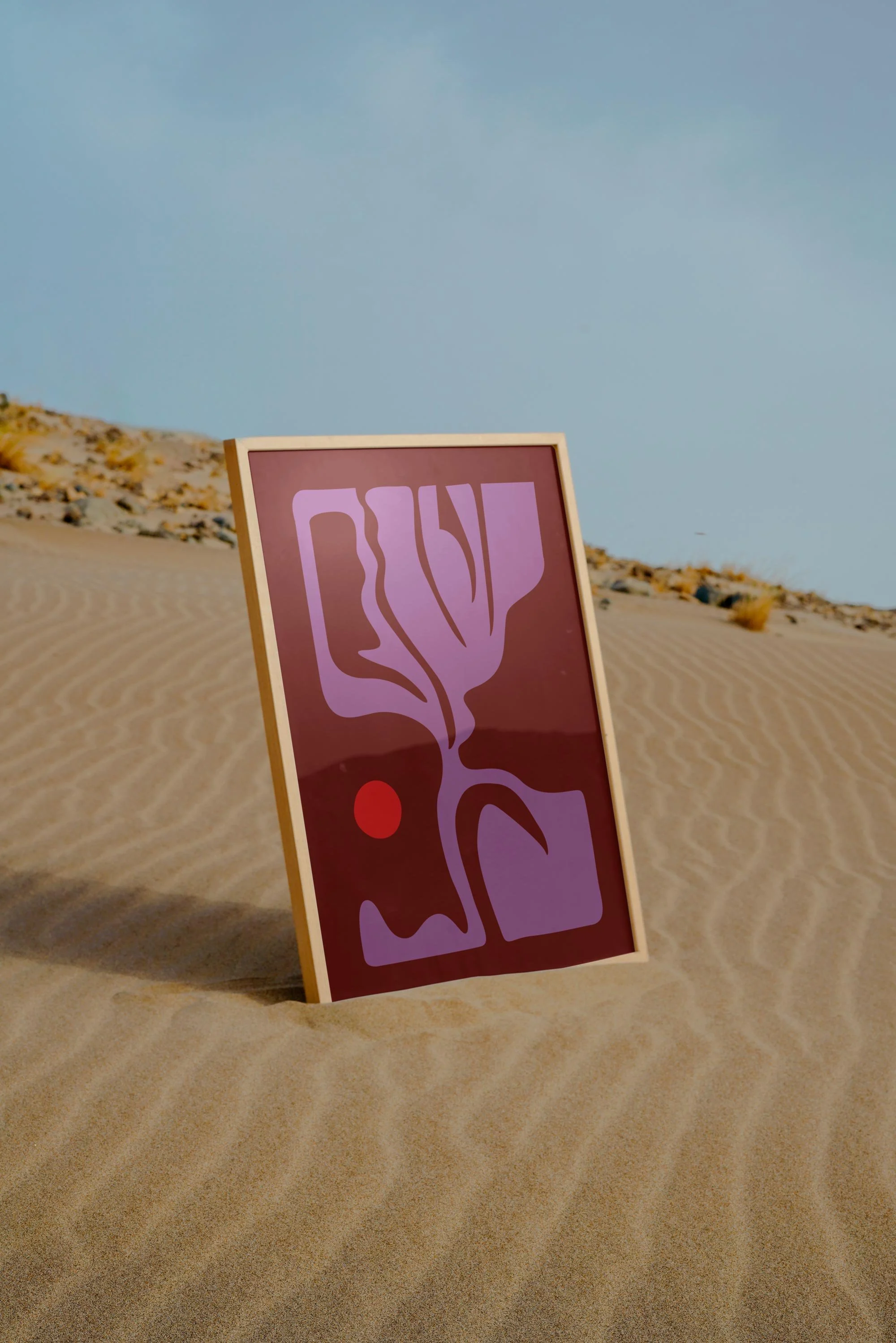 An abstract artwork on a framed canvas, featuring purple and red shapes, placed in a desert landscape with sand dunes under a clear blue sky.