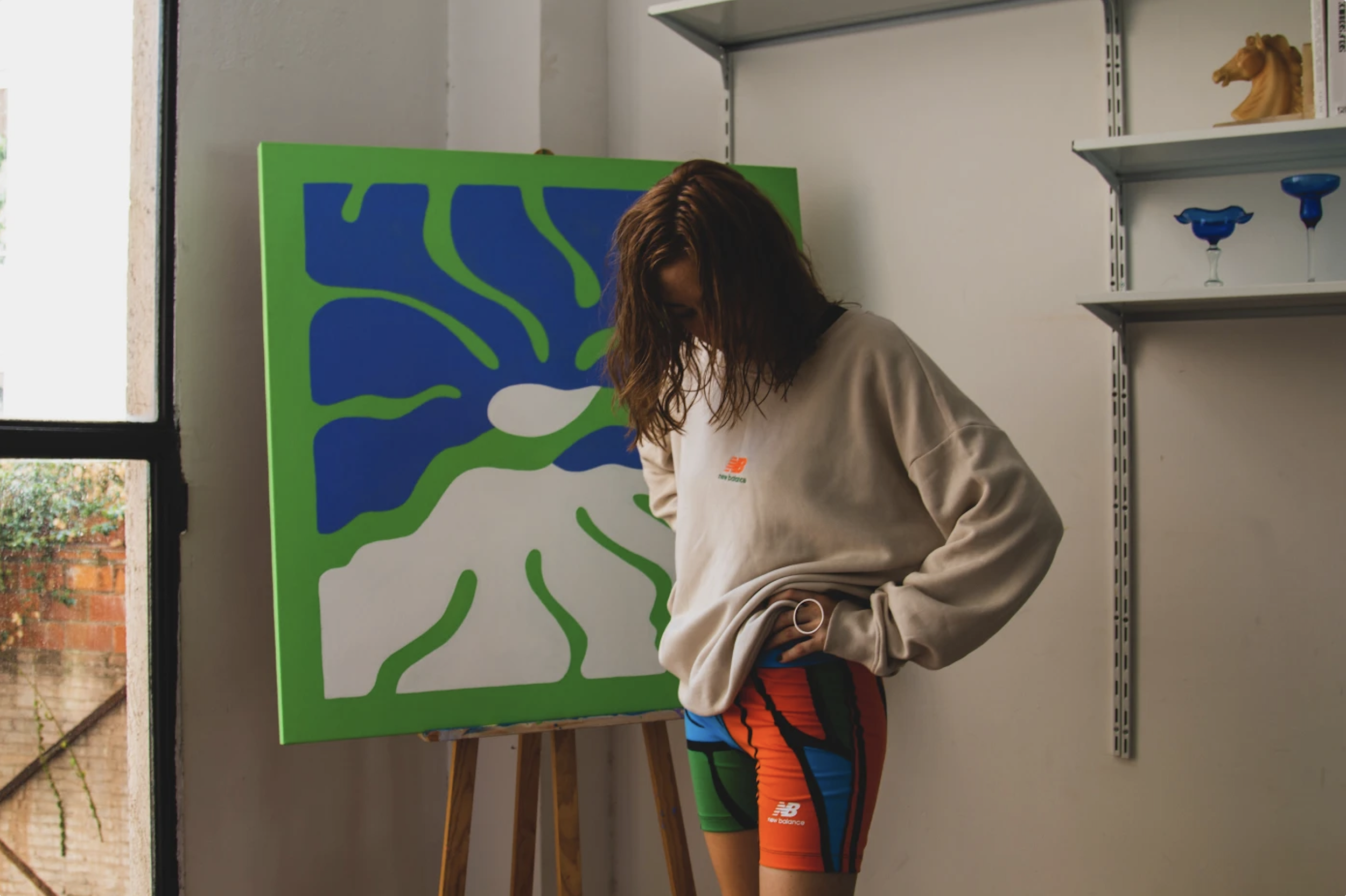 A woman with brown, wavy hair wearing a beige New Balance sweatshirt and colorful athletic shorts, standing in front of a large, abstract, blue and green painting on an easel in an indoor art studio.