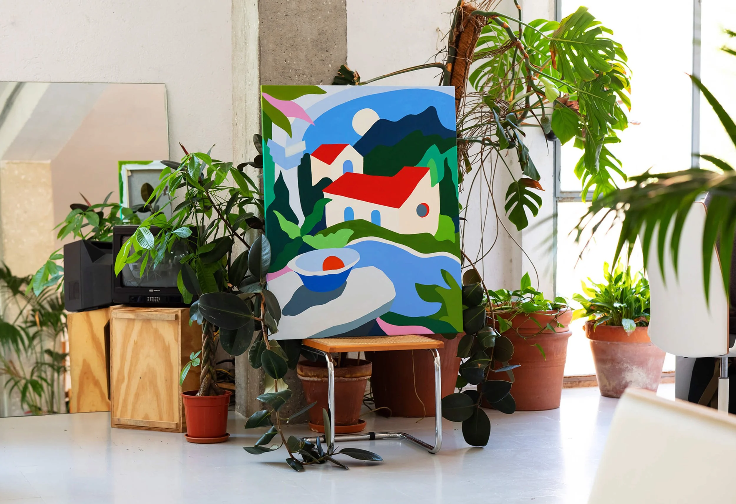 Colorful abstract painting of a house near a river with mountains and a sky in the background, surrounded by green plants in pots inside a room with a light-colored floor and large window.