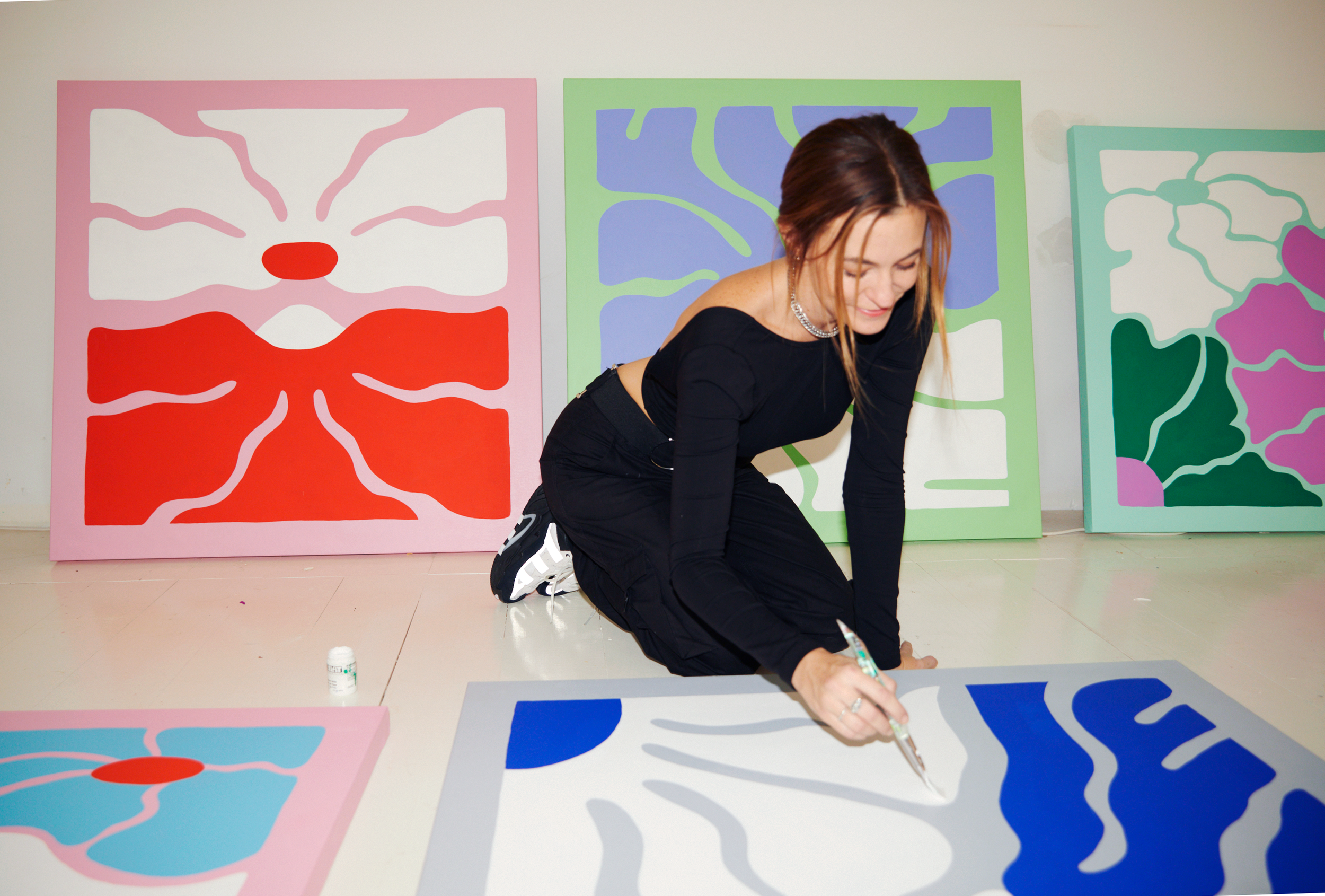 Artist kneeling on the floor, painting abstract artwork on a canvas, with colorful paintings of abstract shapes behind her.