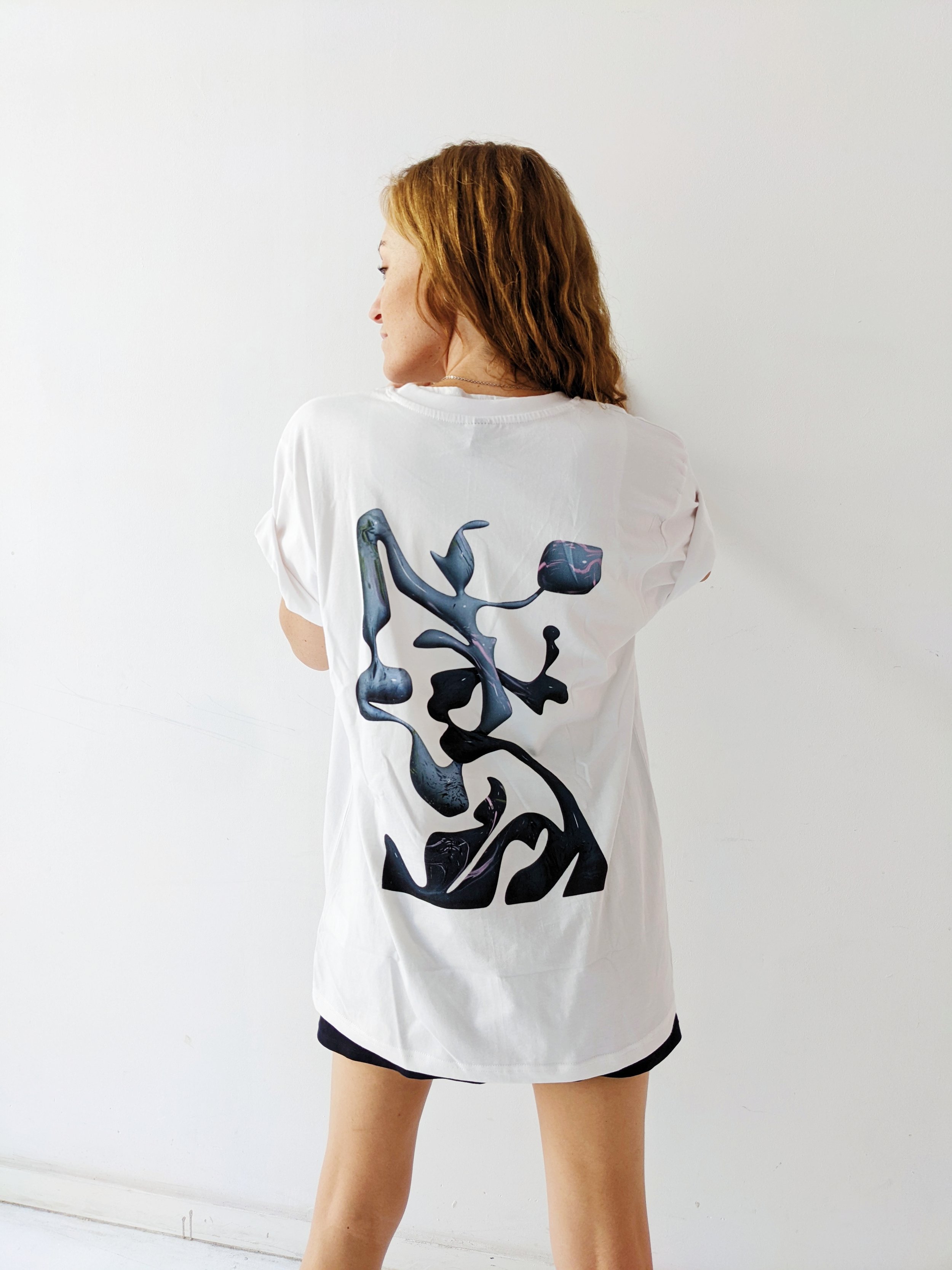 Back view of a woman with reddish hair wearing a white t-shirt with an abstract black and colorful design on the back, standing against a plain white wall.
