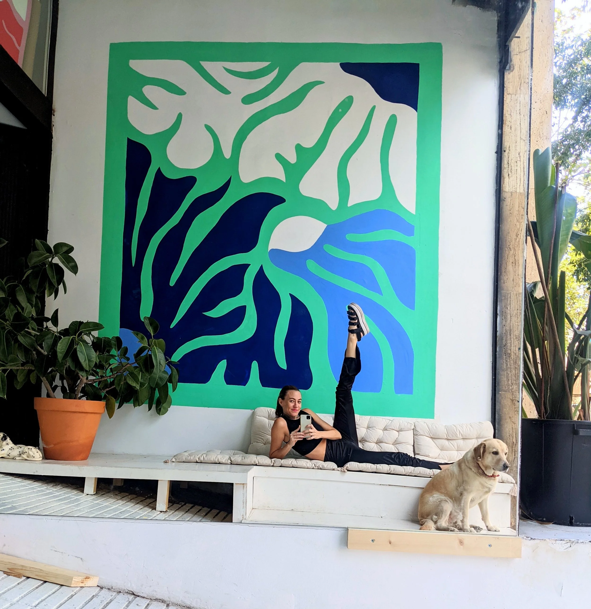 Woman lying on a white cushioned platform taking a selfie, with her leg raised, next to a yellow Labrador sitting on a wooden plank, in front of a blue and green abstract mural, plants in pots on either side.