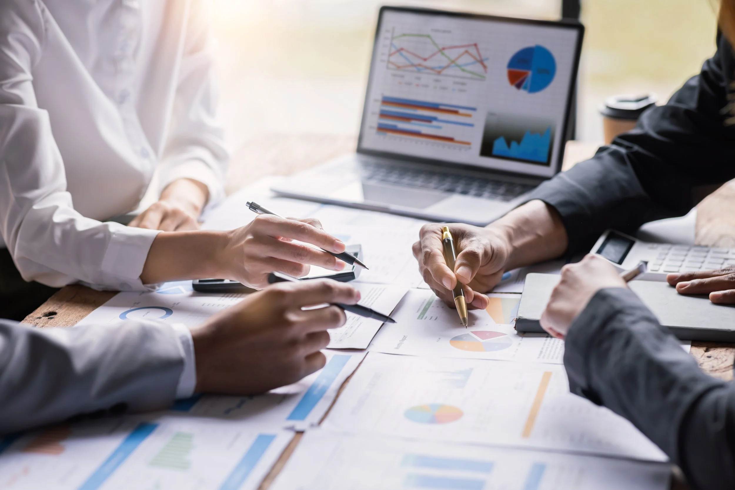 Business people working at a table with documents, charts, and a laptop displaying graphs and pie charts.