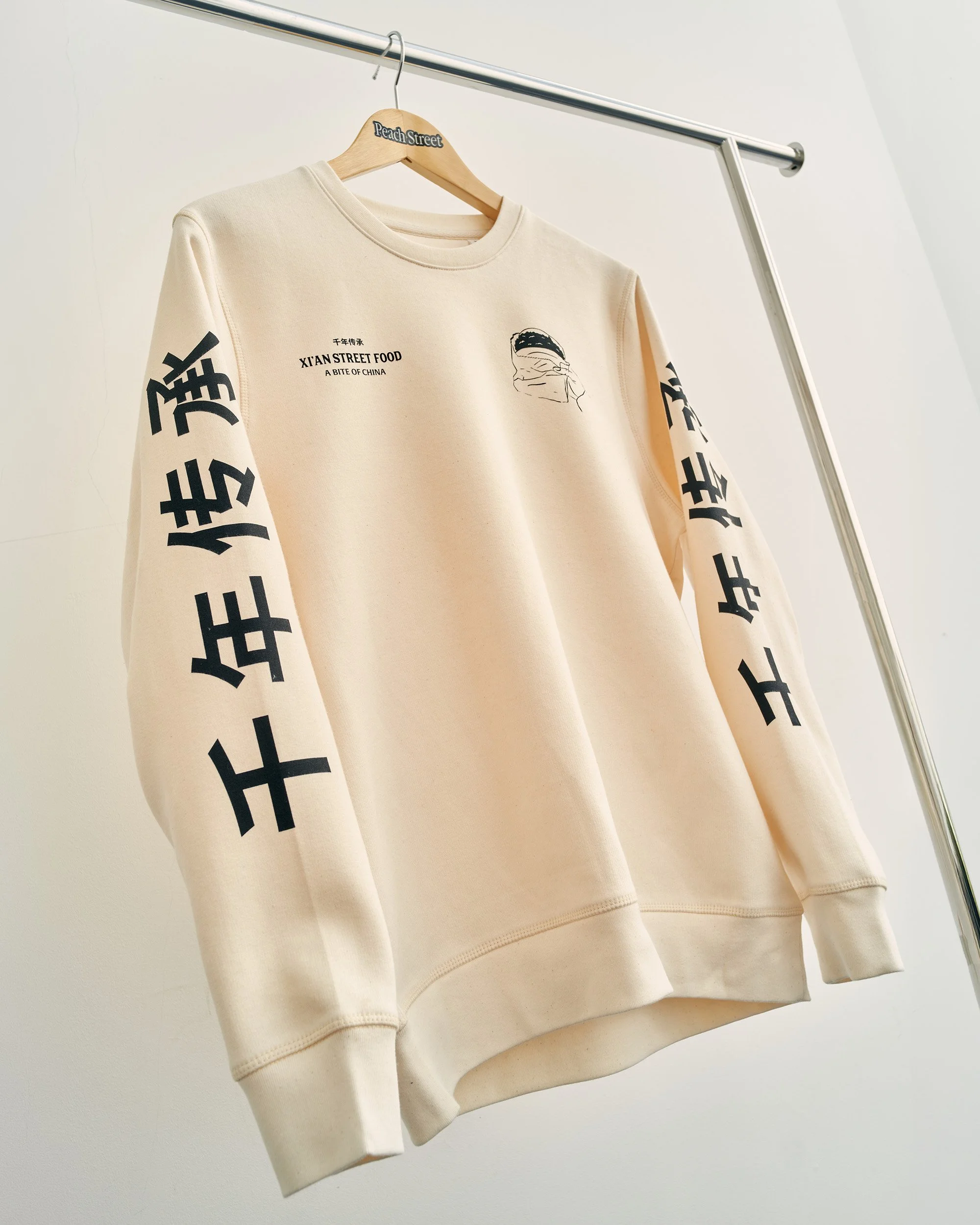 Screen printed by Peach Street for Xian Street Food. A Cream-coloured sweatshirt hanging on a wooden and metal hanger, with black text and graphic designs, on a clothing rack against a white wall.