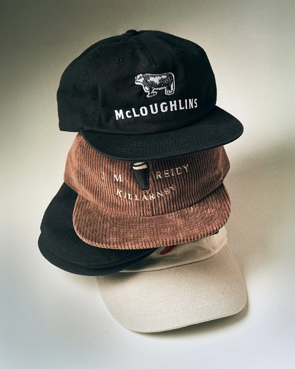 Stack of various baseball caps embroidered by Peach Street, including black with embroidered cow and "McLoughlins" text, brown corduroy with embroidered text, and beige cap, on a plain surface.