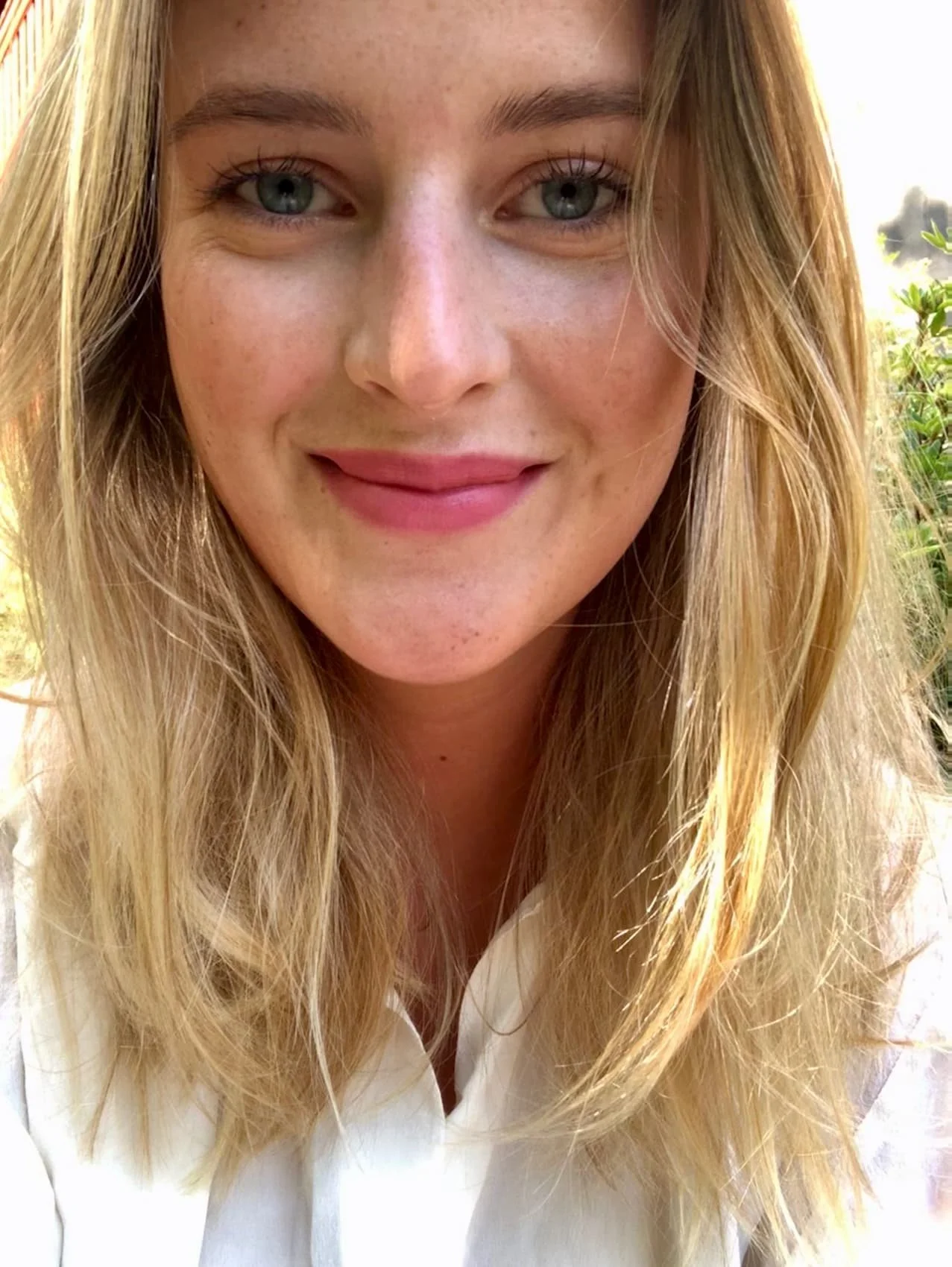Close-up of a smiling woman with blonde hair, blue eyes, wearing a white top, outdoors with greenery in the background.