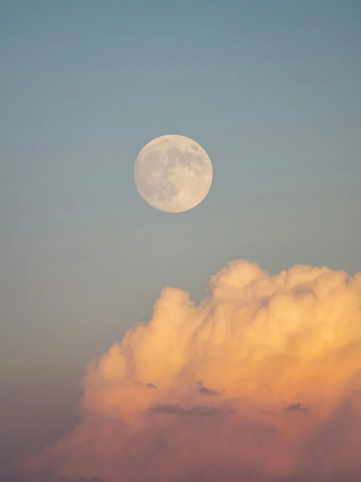 A full moon in the sky above orange and pink clouds at sunset or sunrise.