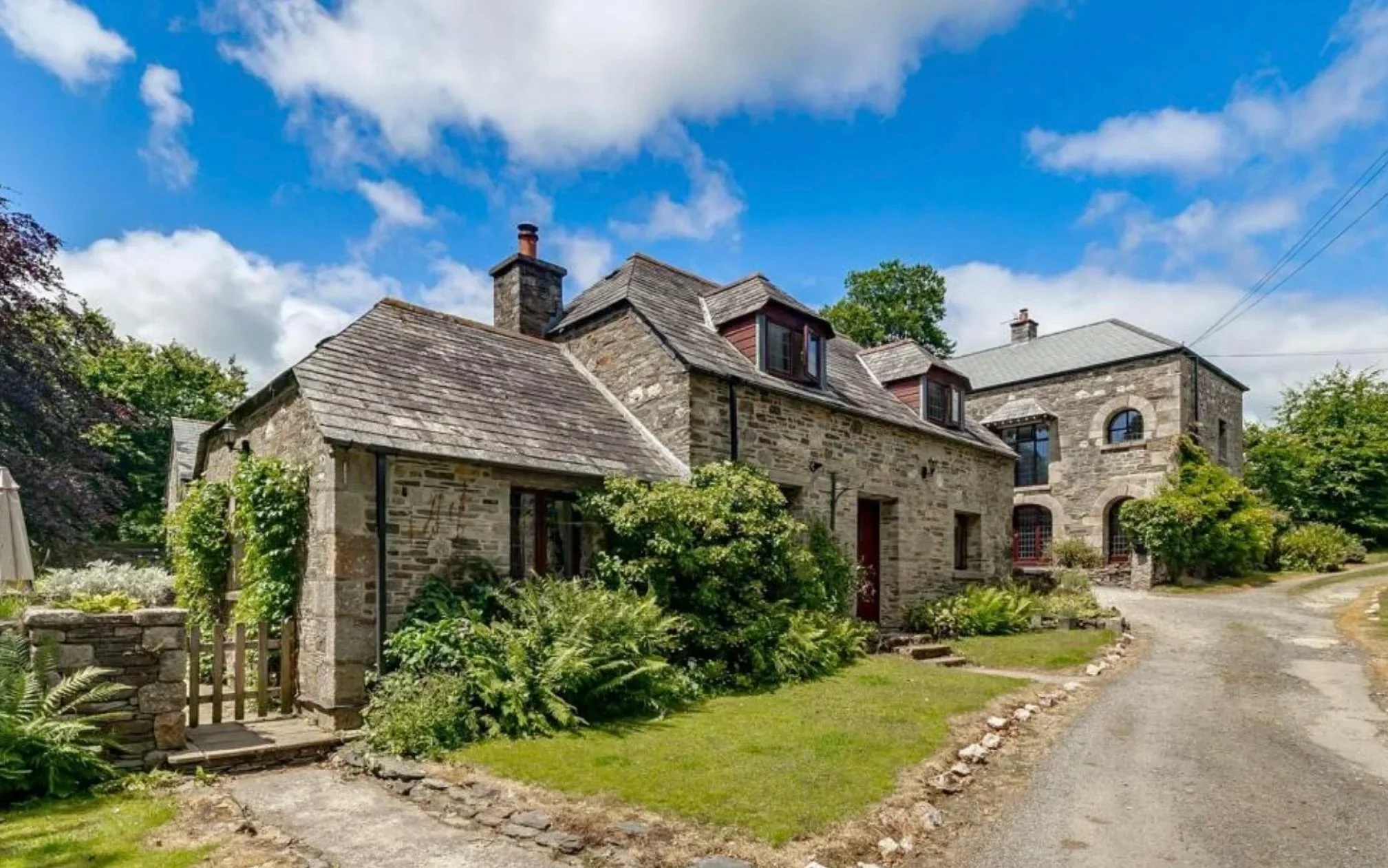 A stone cottage with a garden, trees, and a gravel driveway under a blue sky with clouds.