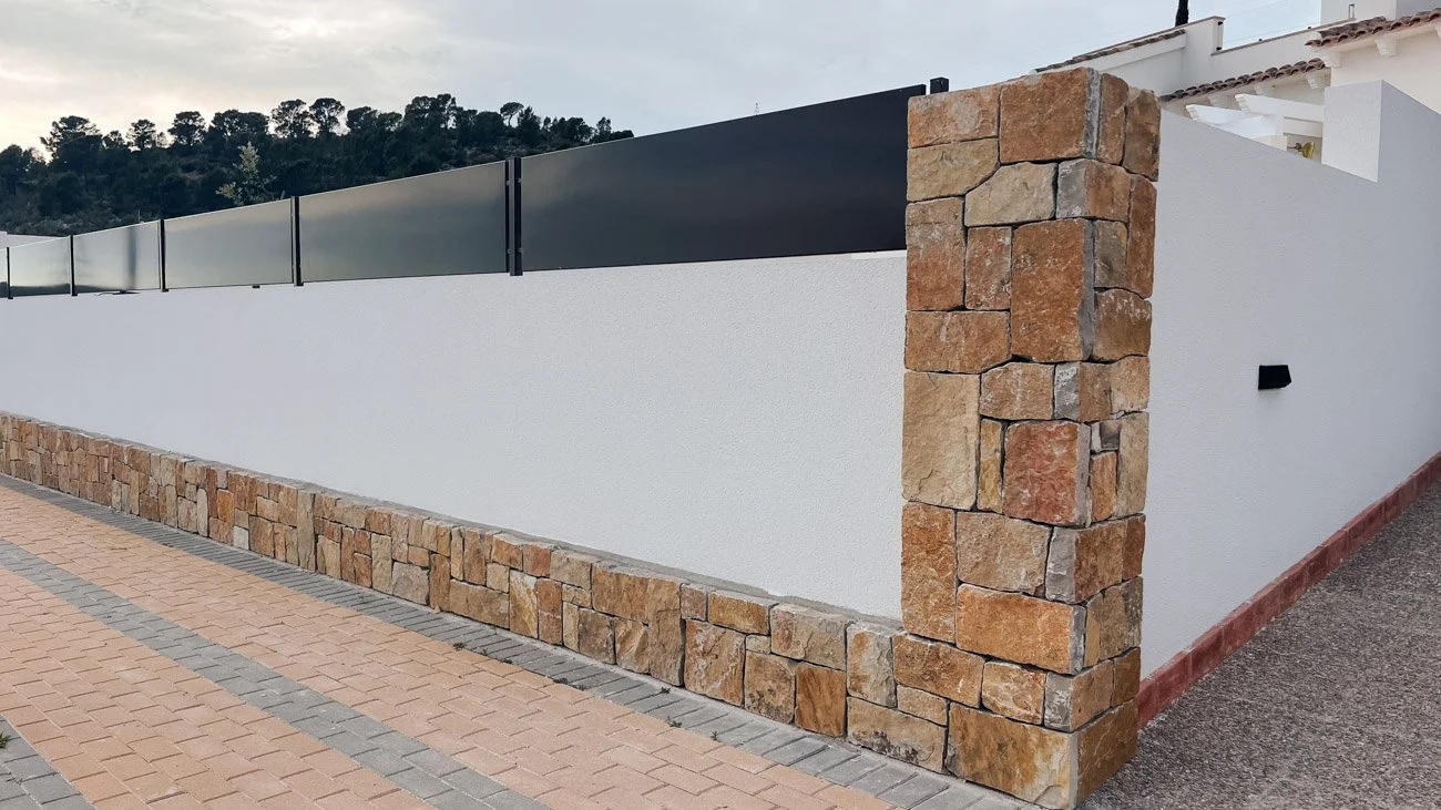 Muro decorativo en la entrada de una casa con bloques de piedra en la esquina y un muro blanco con malla metálica en la cima, en un entorno residencial.