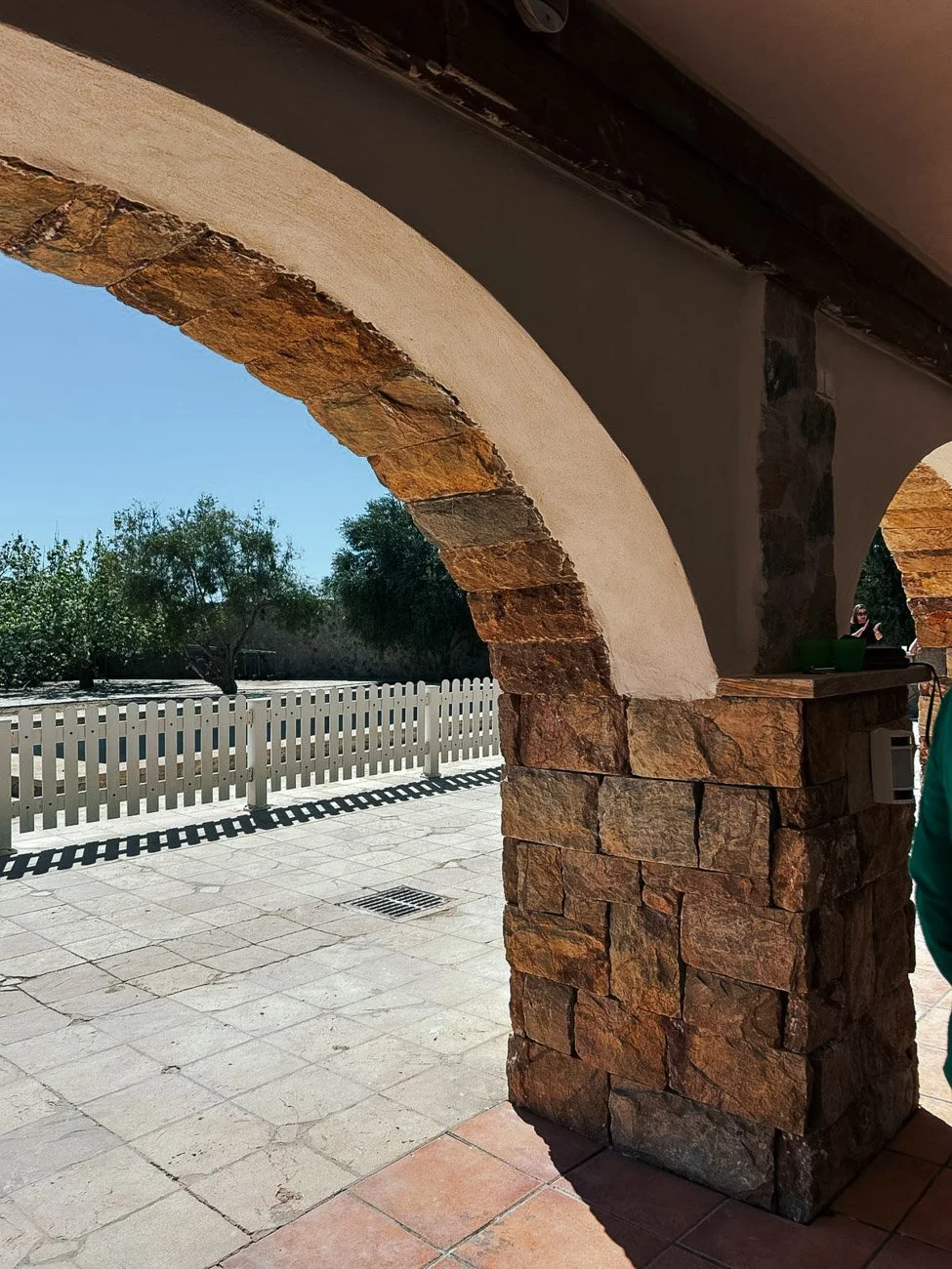 Arcos de piedra y concreto en una estructura exterior con vista a árboles y un cielo despejado, con una cerca de madera blanca y sombra en el suelo.