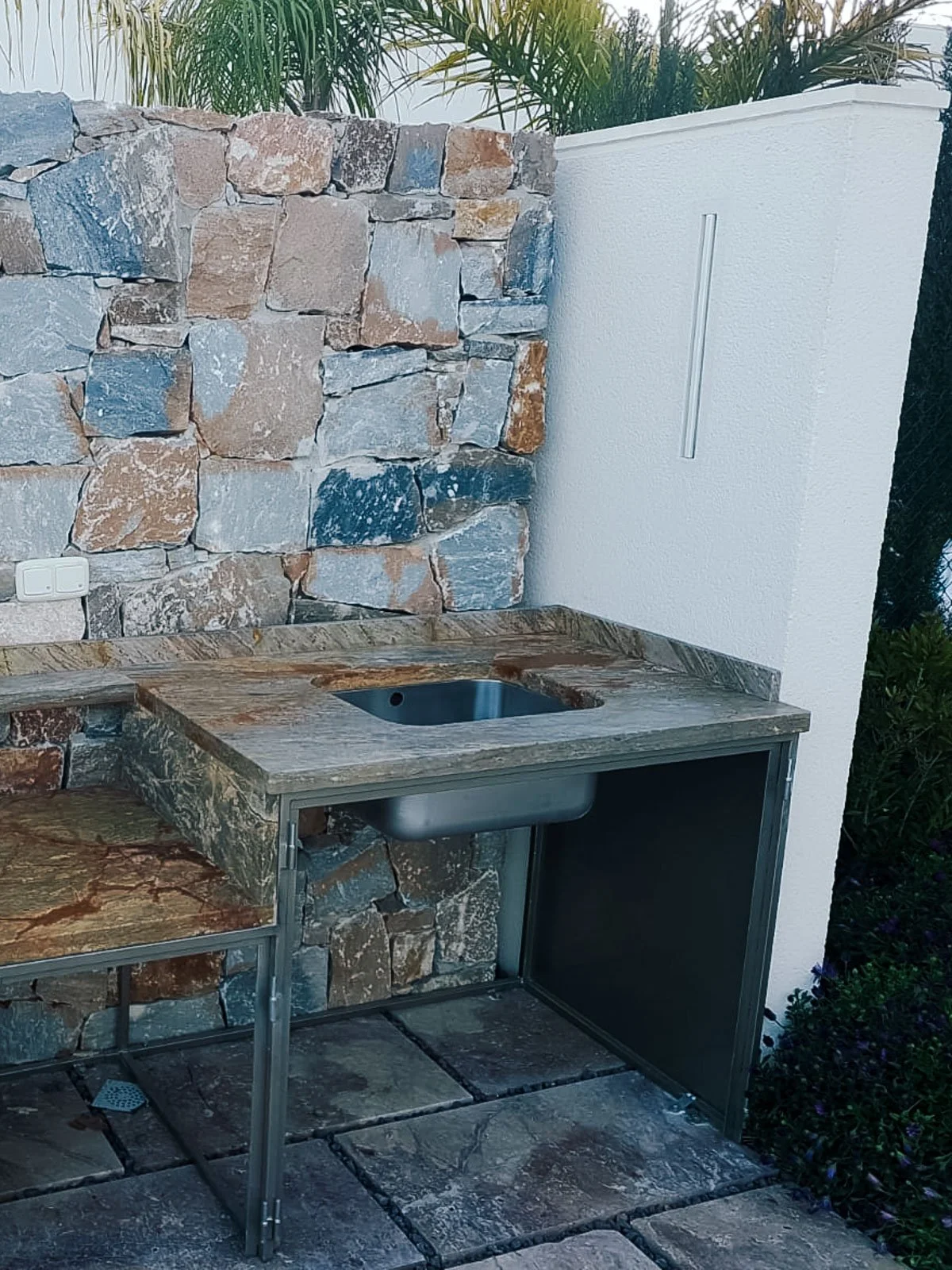 Área exterior con pared de piedra y una pequeña encimera de piedra con un fregadero de metal, rodeada de plantas.