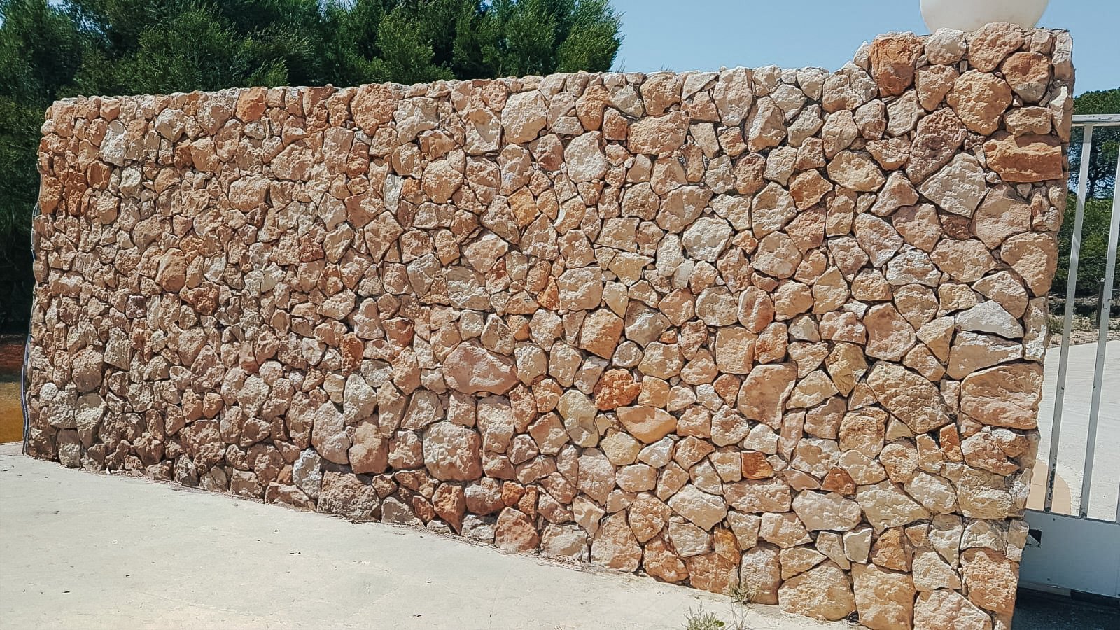 Muro de piedra de color beige y marrón, construido con rocas irregulares y tamaño variado, en un entorno exterior con árboles y cielo azul.
