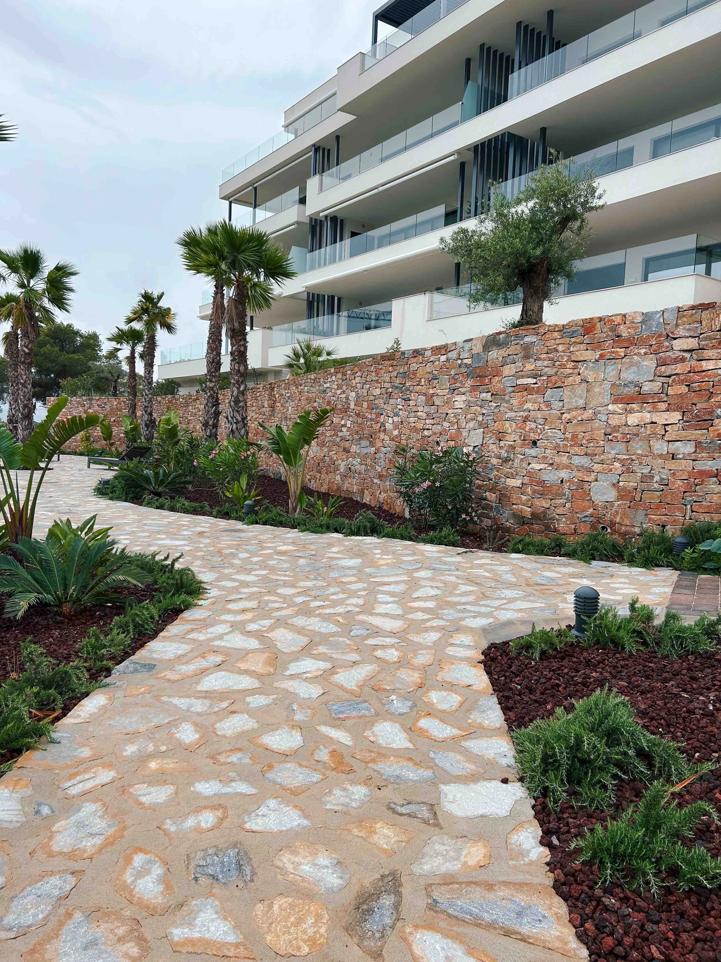 Camino de piedra rodeado de plantas y palmeras, con edificio moderno de apartamentos en el fondo y muro de piedra.