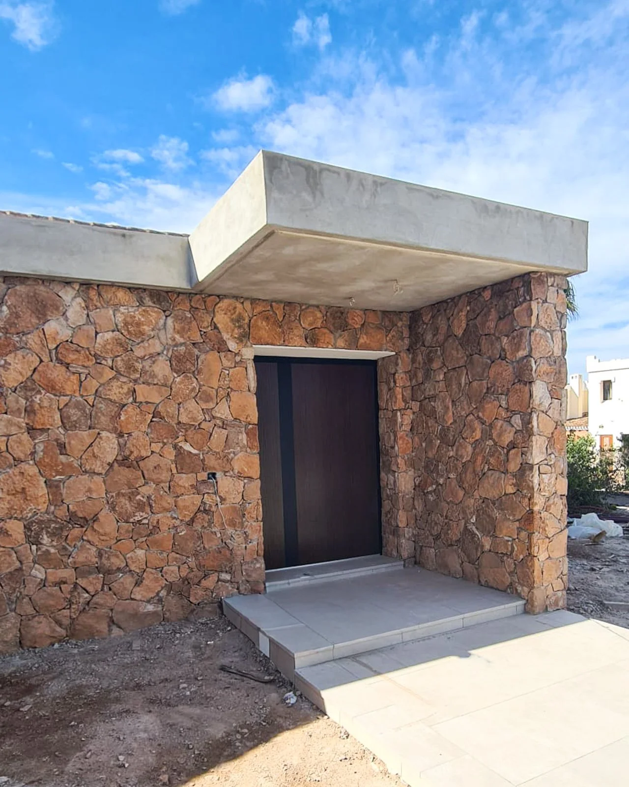 Fachada de casa con muro de piedra y puerta negra, entrada con escalón de piedra y sobrevuelo de concreto, cielo despejado.