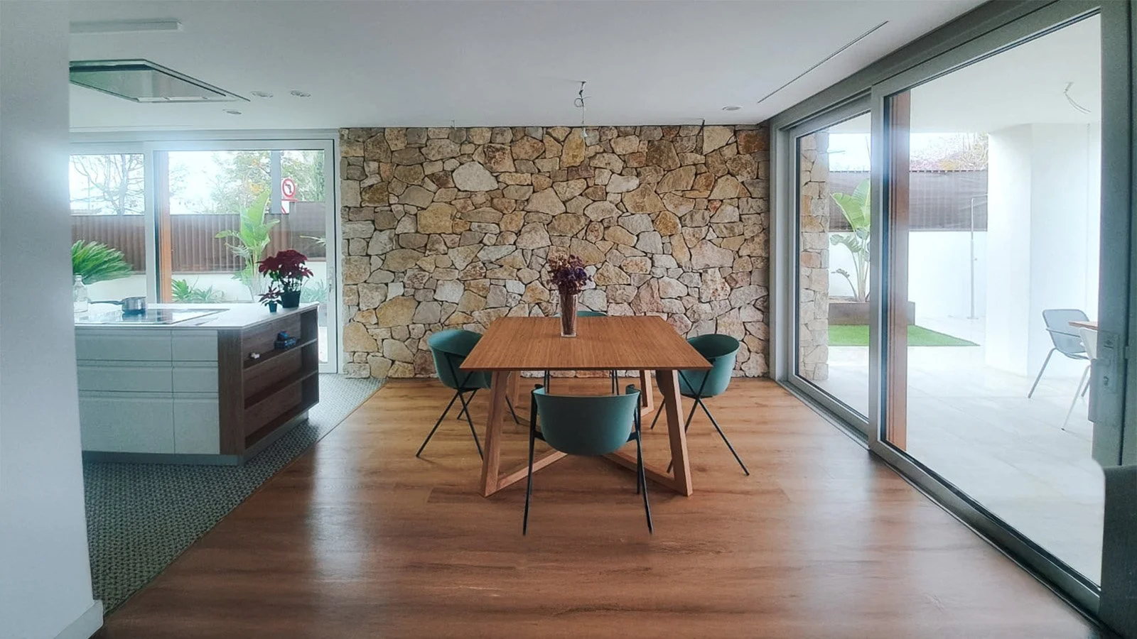 Comedor con mesa de madera y cuatro sillas verdes, pared de piedra en el fondo y ventanas grandes con vista a un jardín exterior.