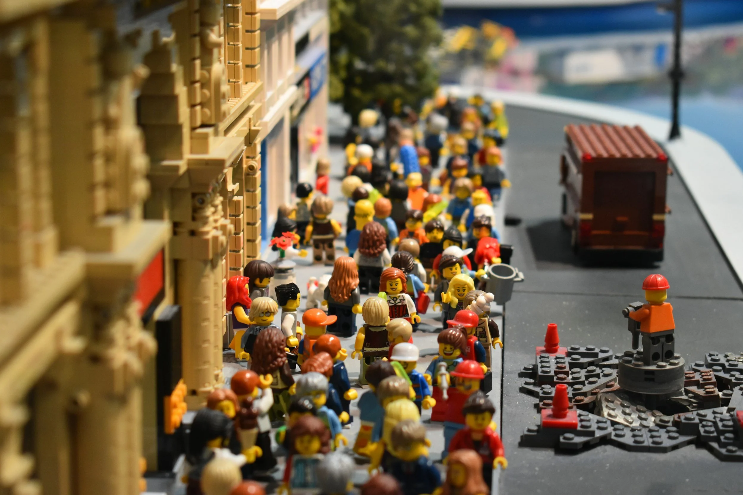 Lego minifigures gathered along a sidewalk in front of a building, with a street and a small truck in the background.