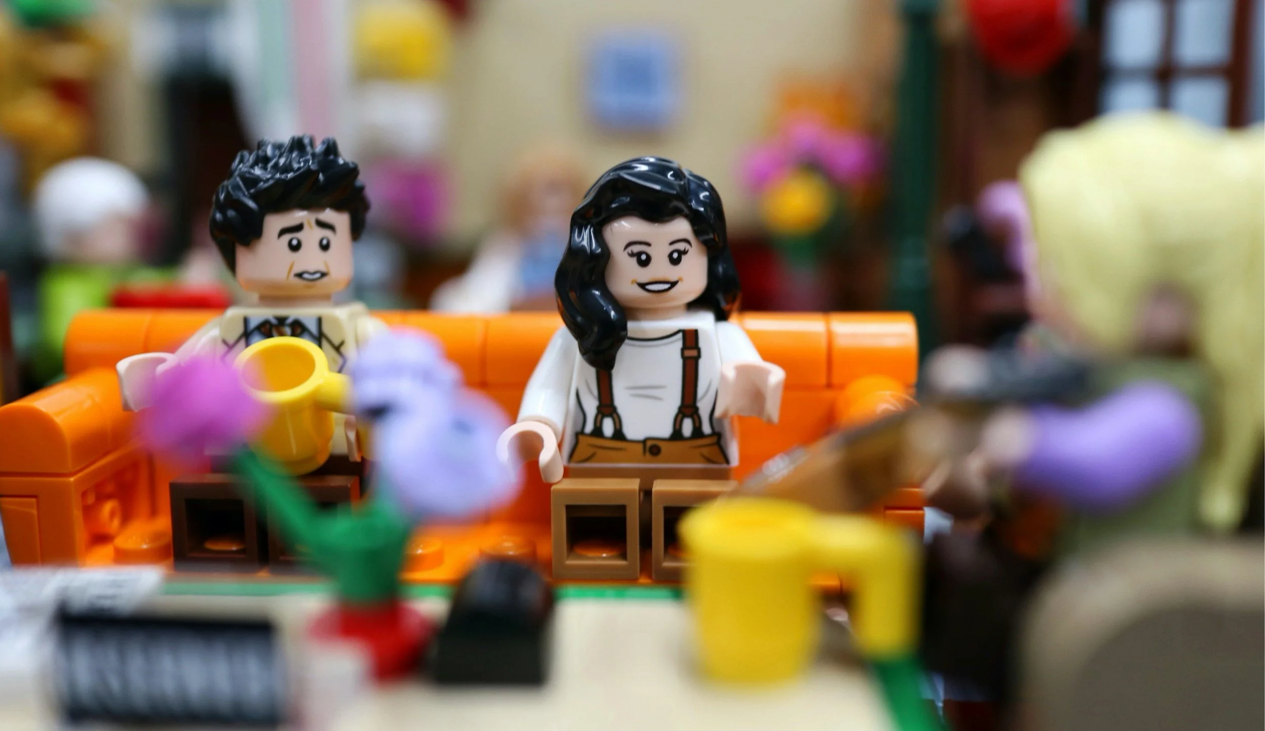 LEGO figurines of a man and a woman sitting on an orange sofa, with various LEGO pieces and flowers in the foreground, and a blurred background of colorful decor.