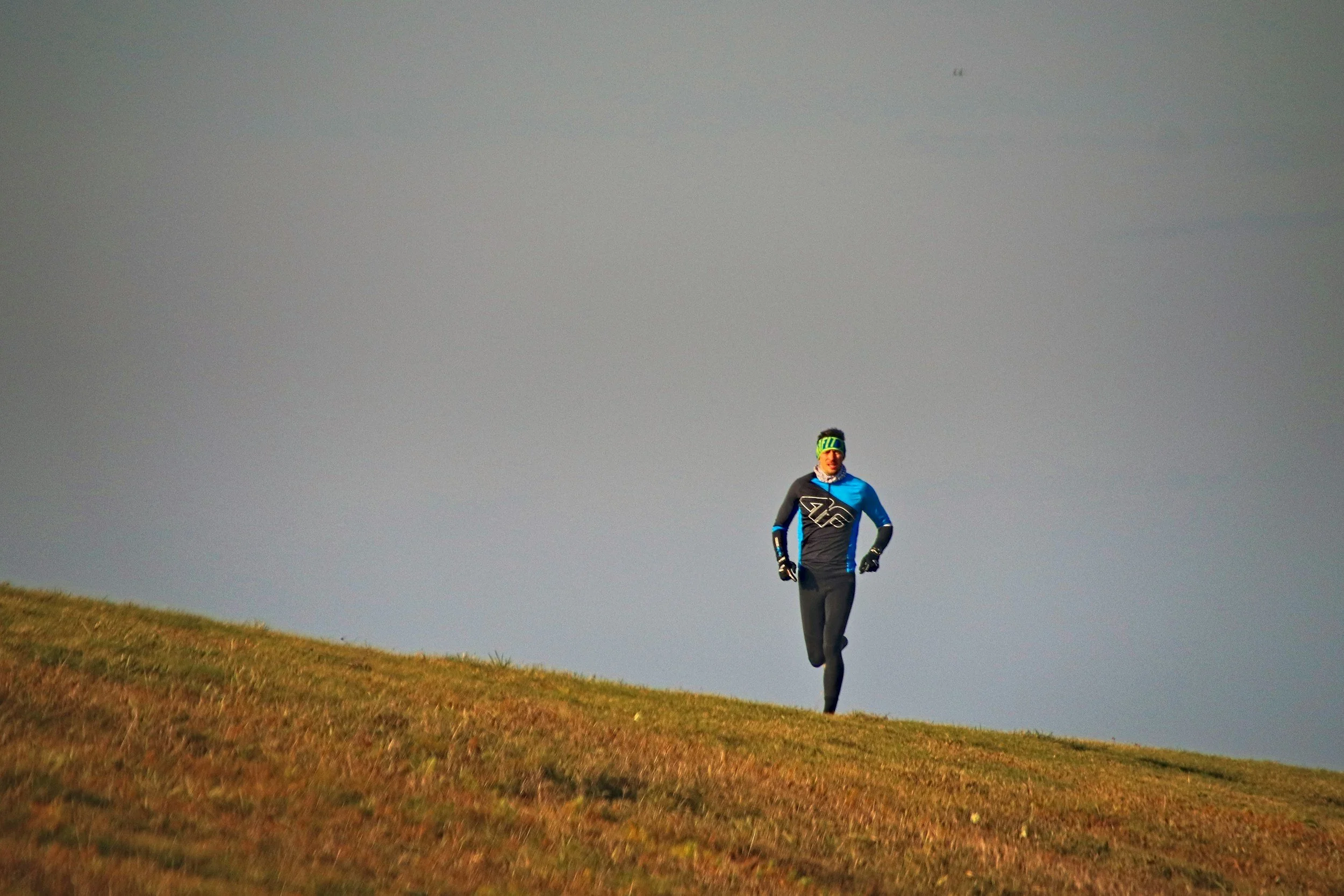 A person dressed in blue and black athletic gear running uphill on a grassy hill, with a cloudy sky in the background.