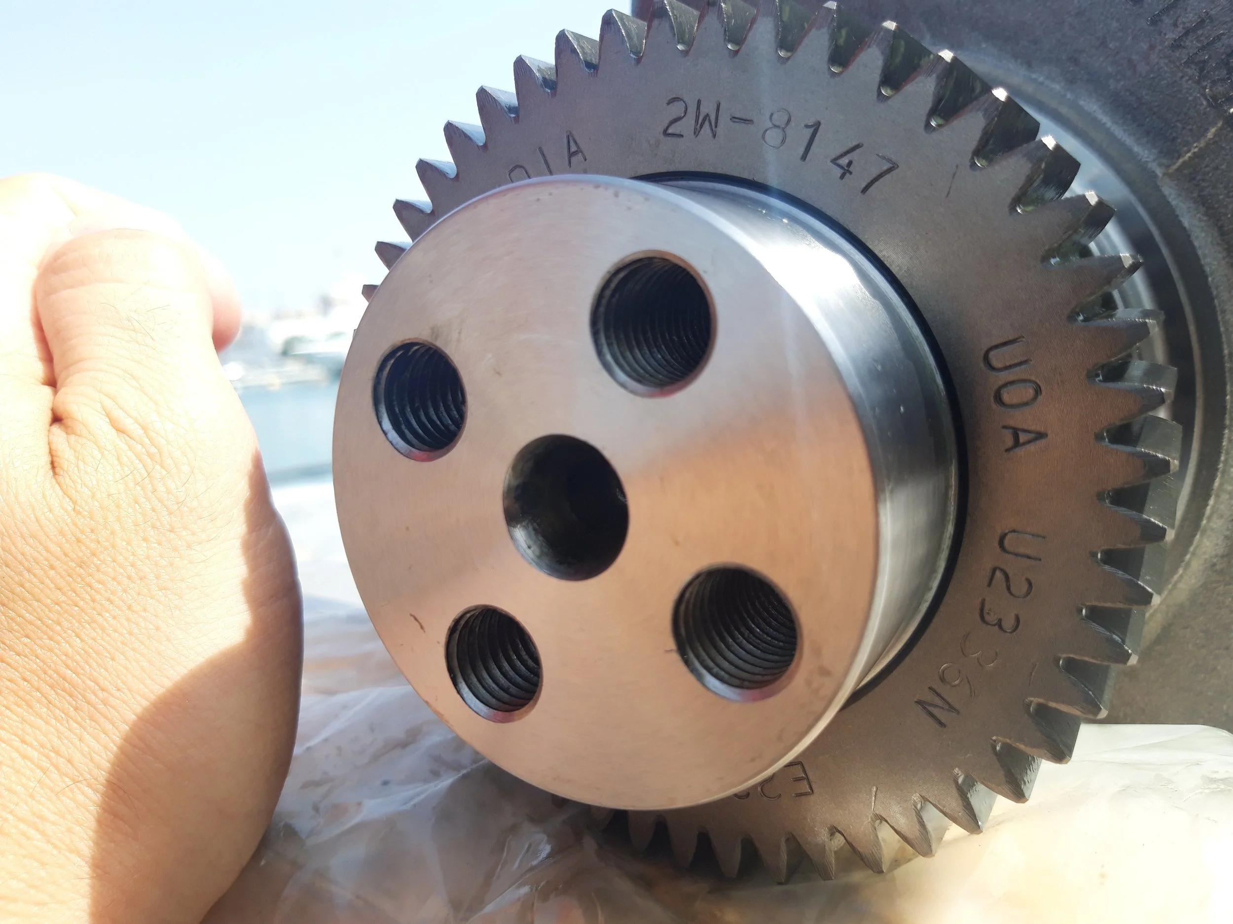 Close-up of a large metal gear or cogwheel with five threaded holes and engraved markings, held by a person with a hand visible on the left side.