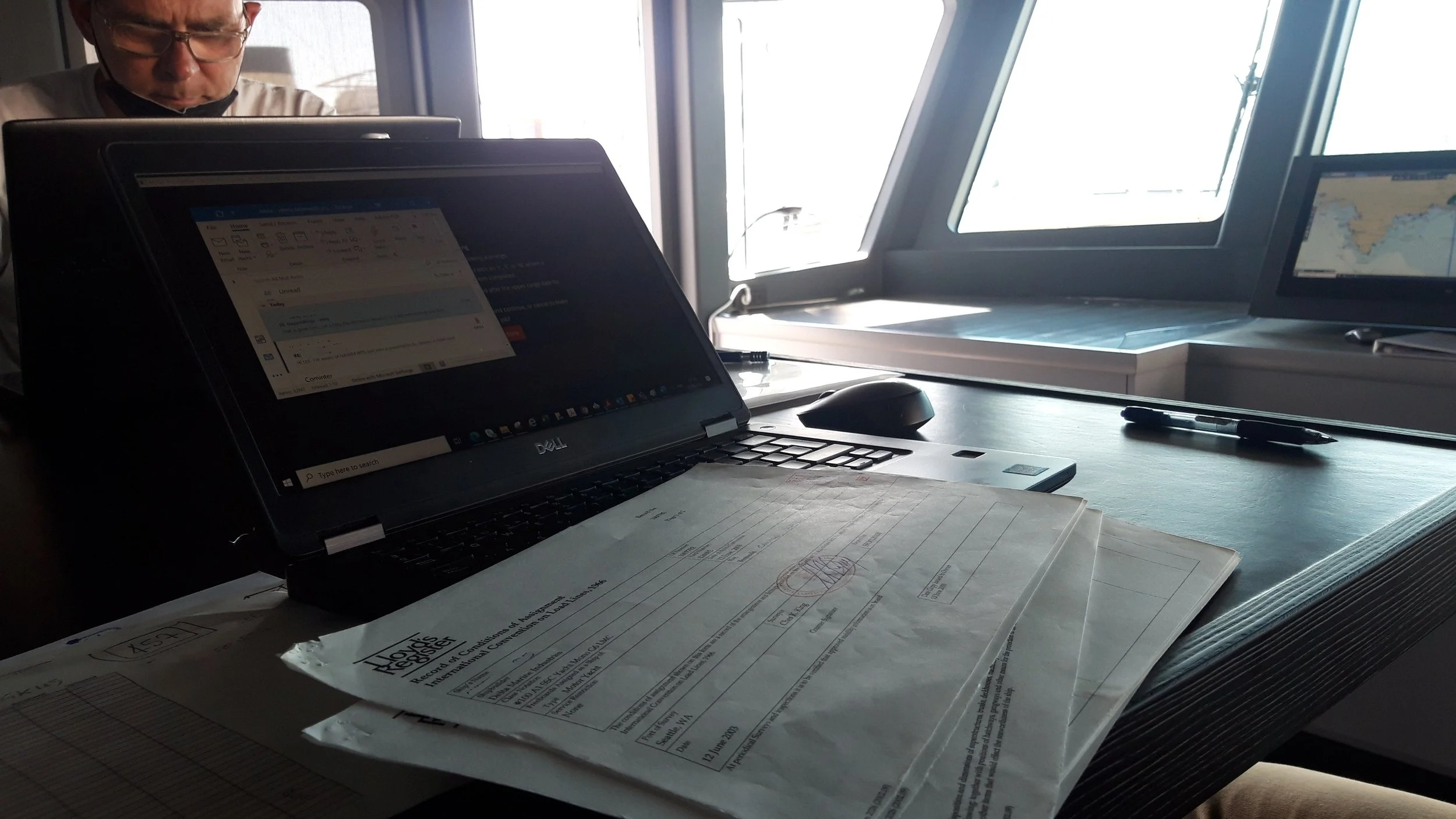 Office desk with a laptop, papers, a pen, a computer mouse, and a window in the background showing bright daylight.