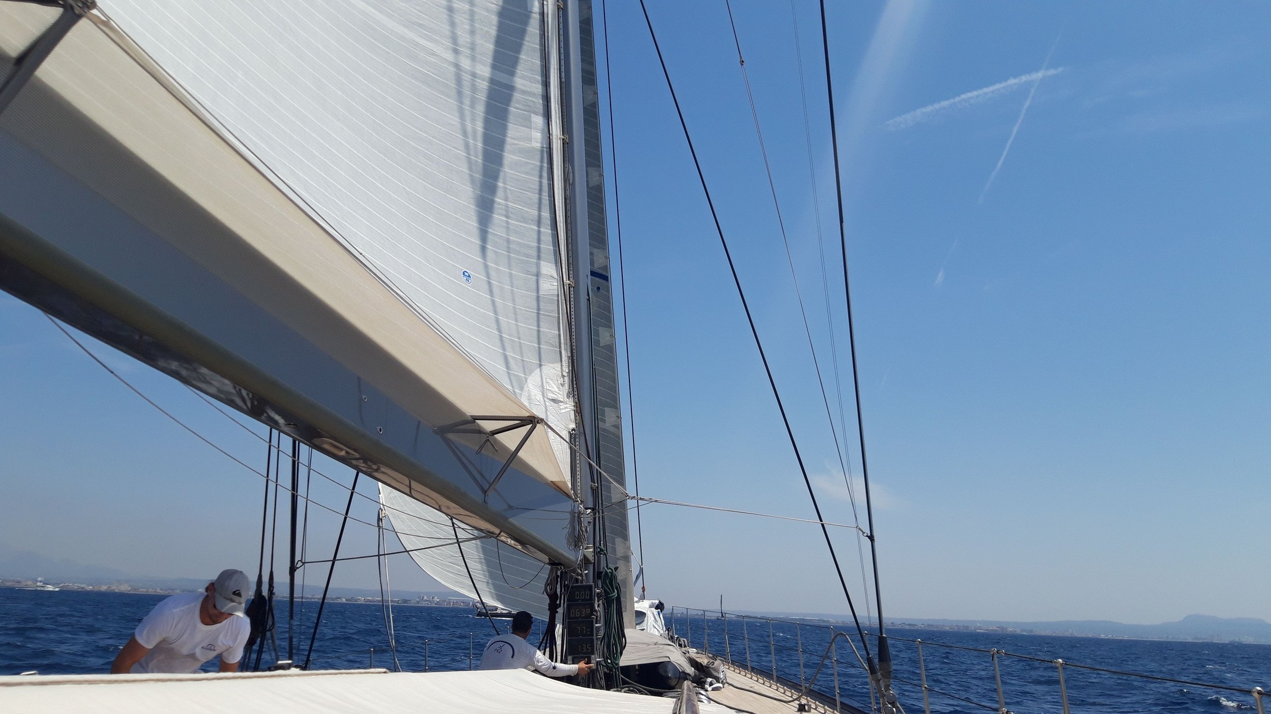 View from a sailing boat showing the sail, rigging, and crew member adjusting equipment against a blue sky and ocean.