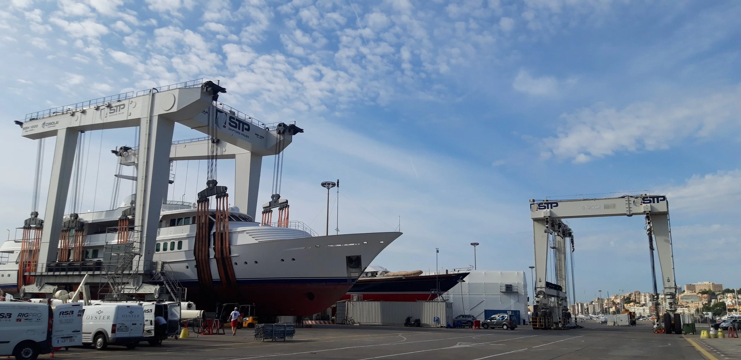 A large yacht is being worked on in a shipyard, lifted out of the water by two large, white gantry cranes marked with the 