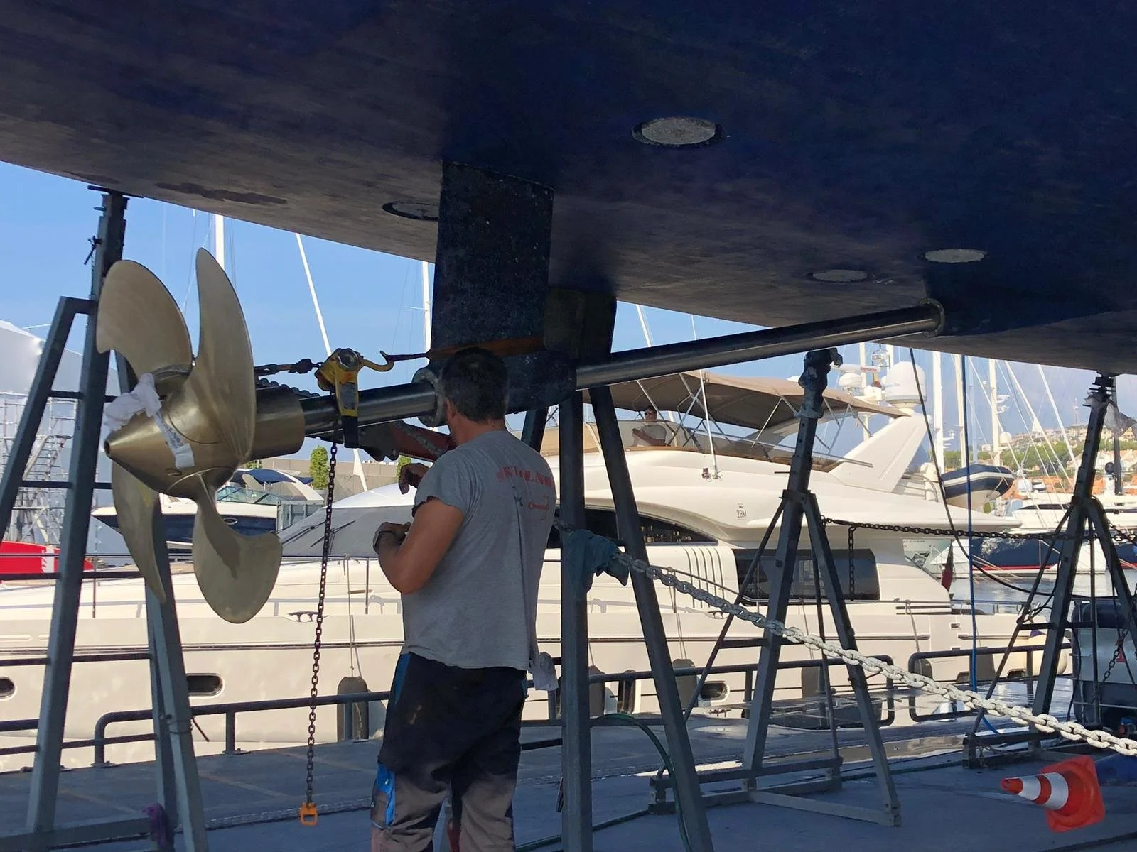A man working on a boat's propeller at a marina, with several docked yachts in the background.