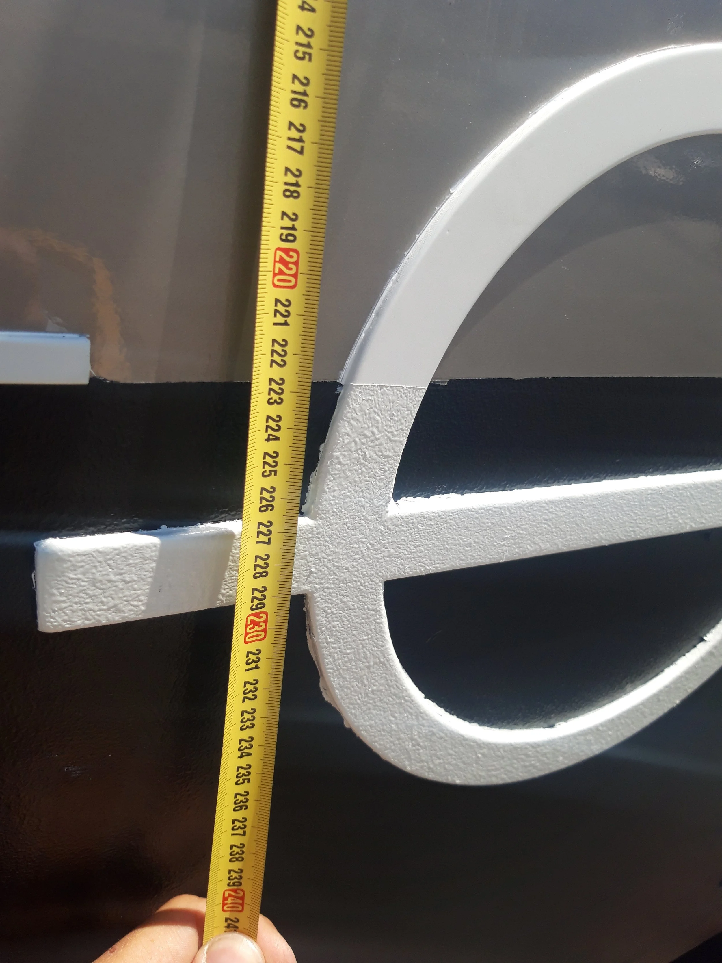 Close-up of a measuring tape showing approximately 220 centimeters, held against a decorative metal sign with a large white letter 'E' on a dark background.