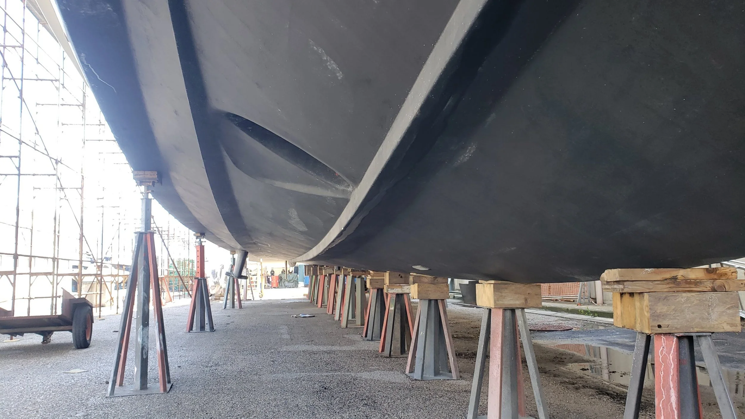 Close-up of a boat under construction, supported by work stands, with scaffolding and construction tools visible in the background.