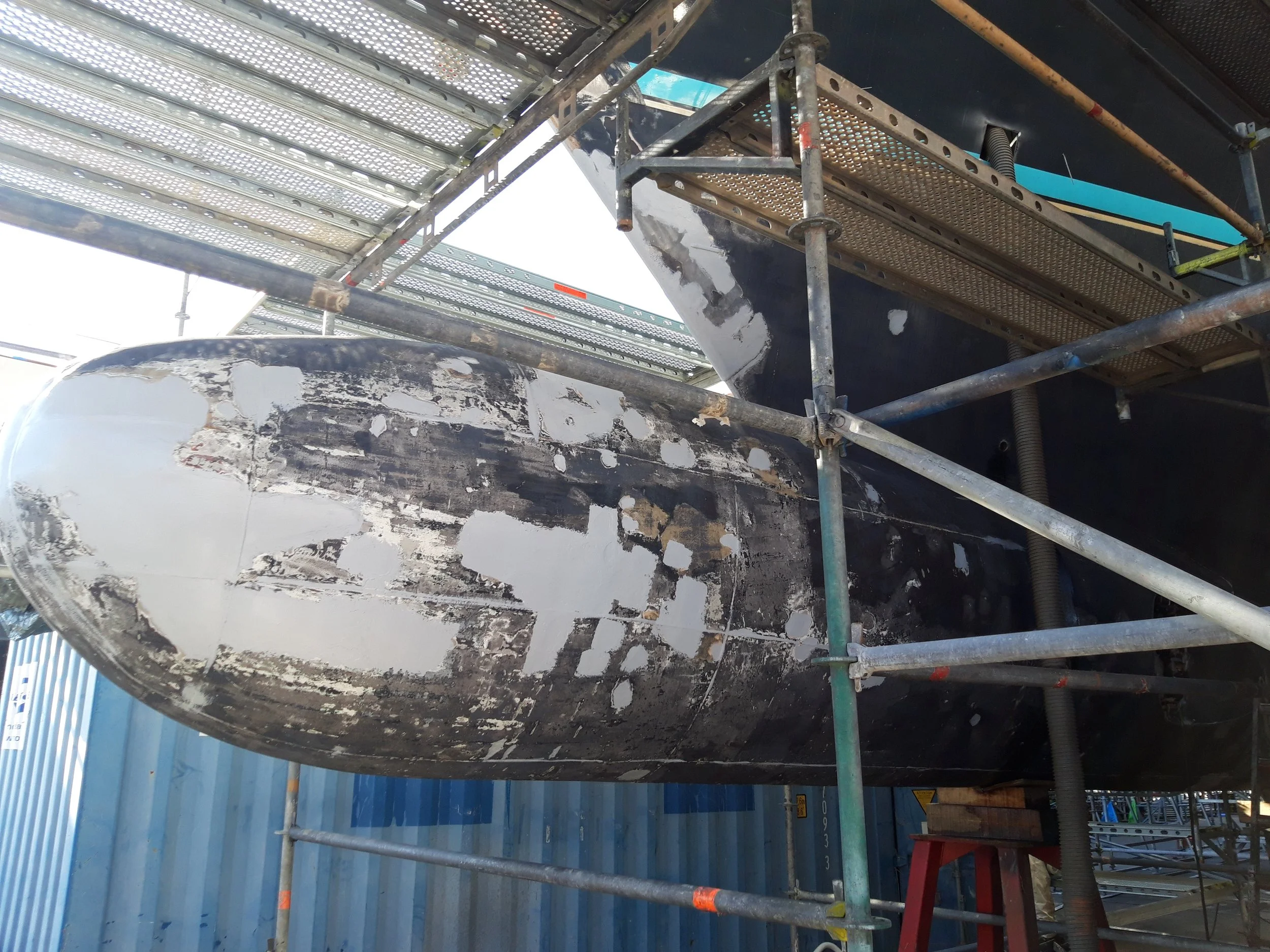 Close-up view of the hull of a boat with peeling paint, supported on scaffolding at a dock, with a blue container in the background.