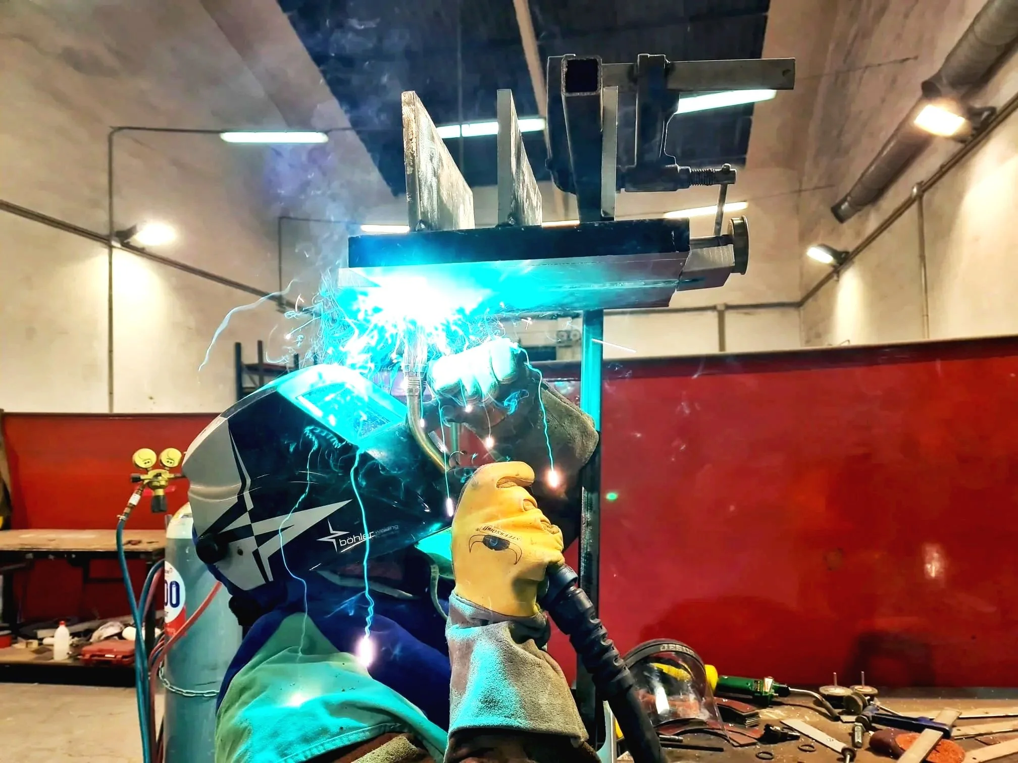 A person welding metal with bright blue sparks inside a workshop, wearing a welding helmet and protective gloves.